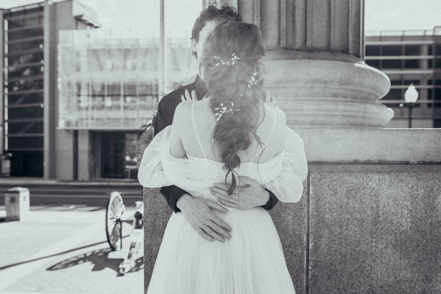 Black and white photo of newlyweds sharing tender embrace beside stone column at outdoor urban venue.