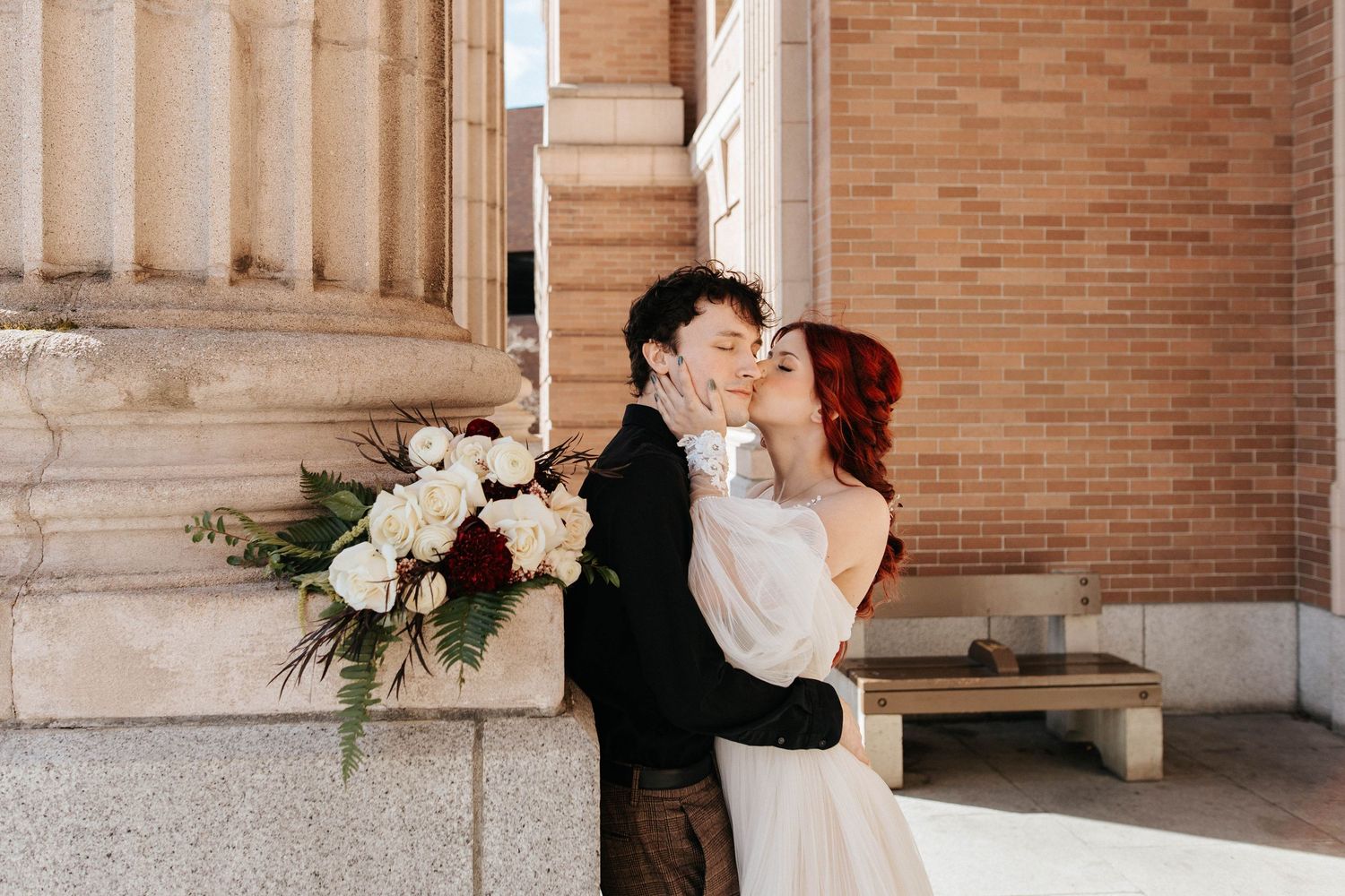Bride and groom sharing a kiss outside a brick building with white floral bouquet nearby.