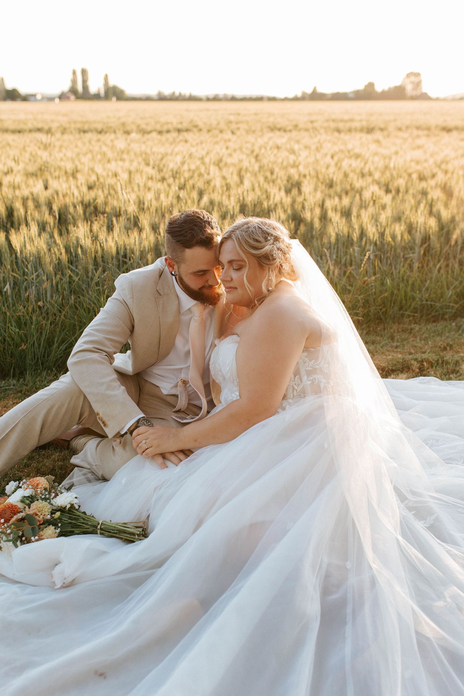Bride and groom sit together in golden wheat field at sunset, sharing an intimate moment in wedding attire.
