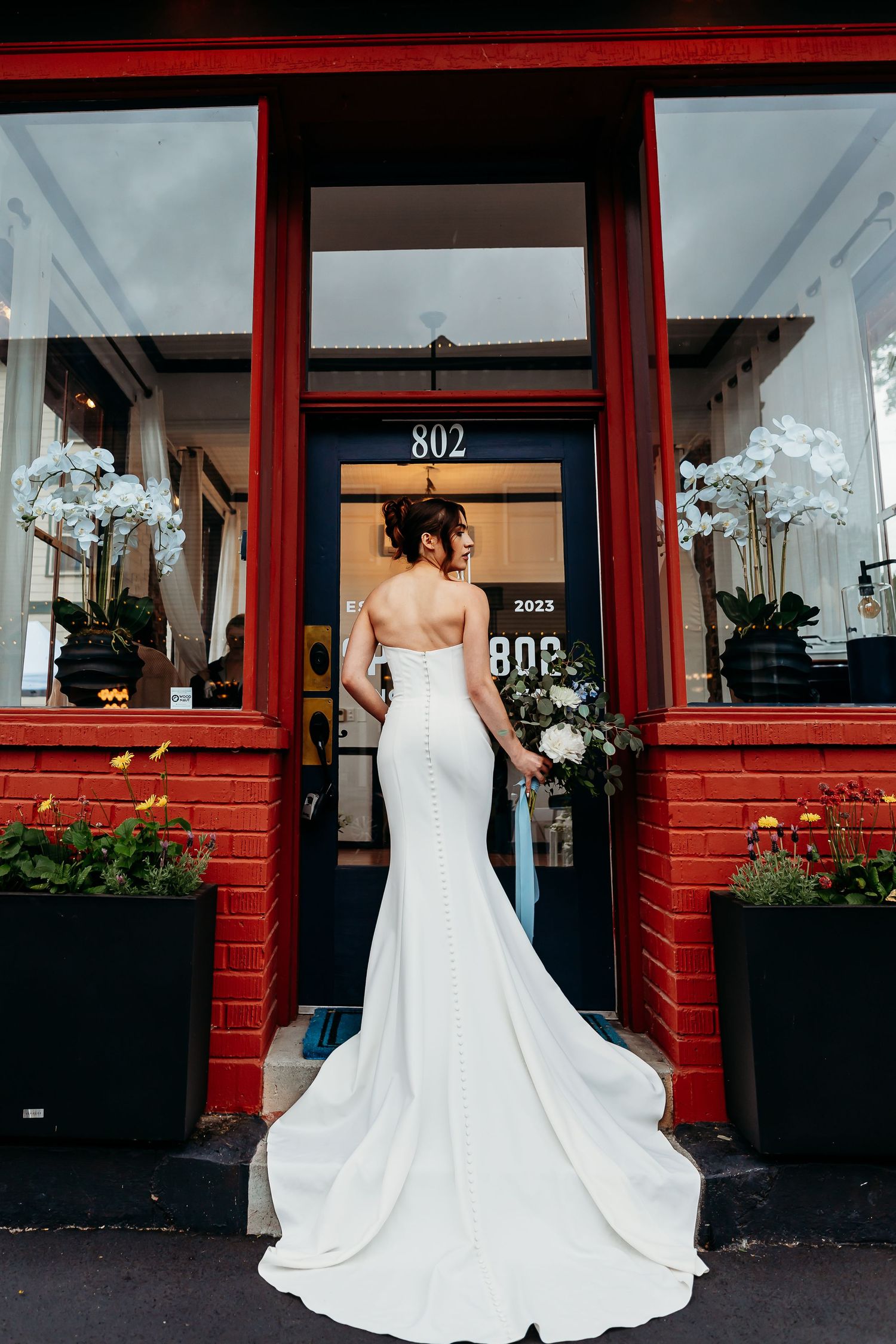 Bride wearing elegant strapless white mermaid gown stands in front of red brick storefront with door number 802.