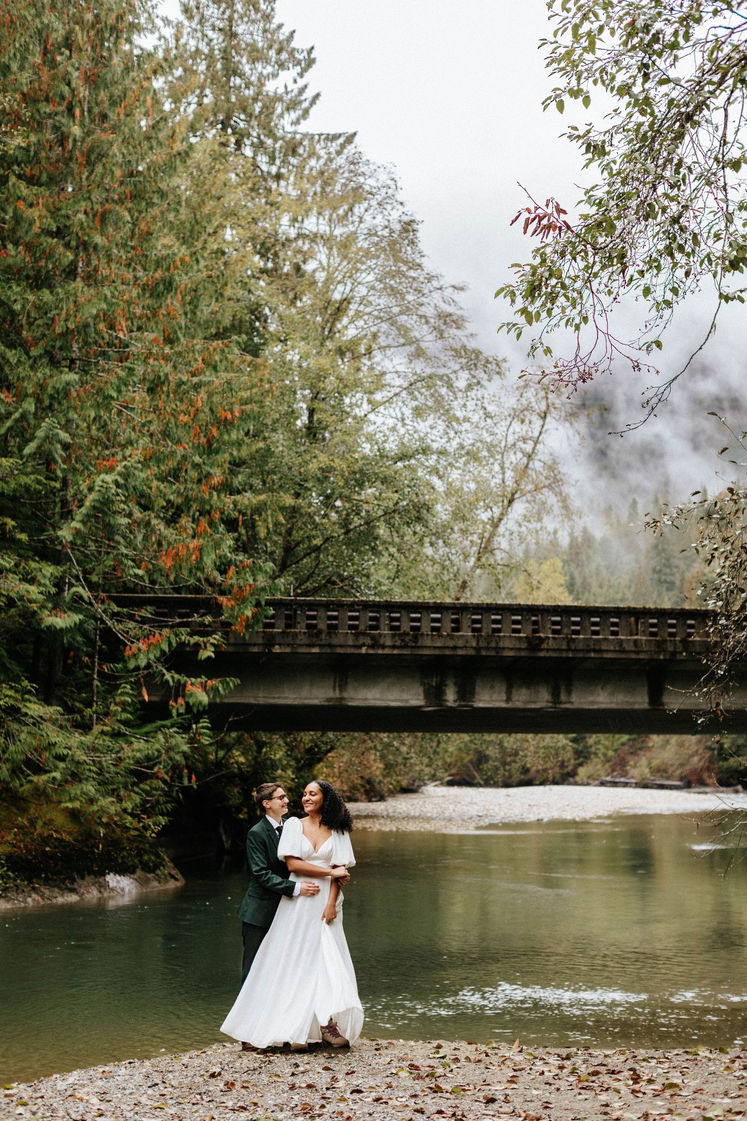 Bride and groom embrace by a misty river with a wooden bridge and autumn trees in the background.