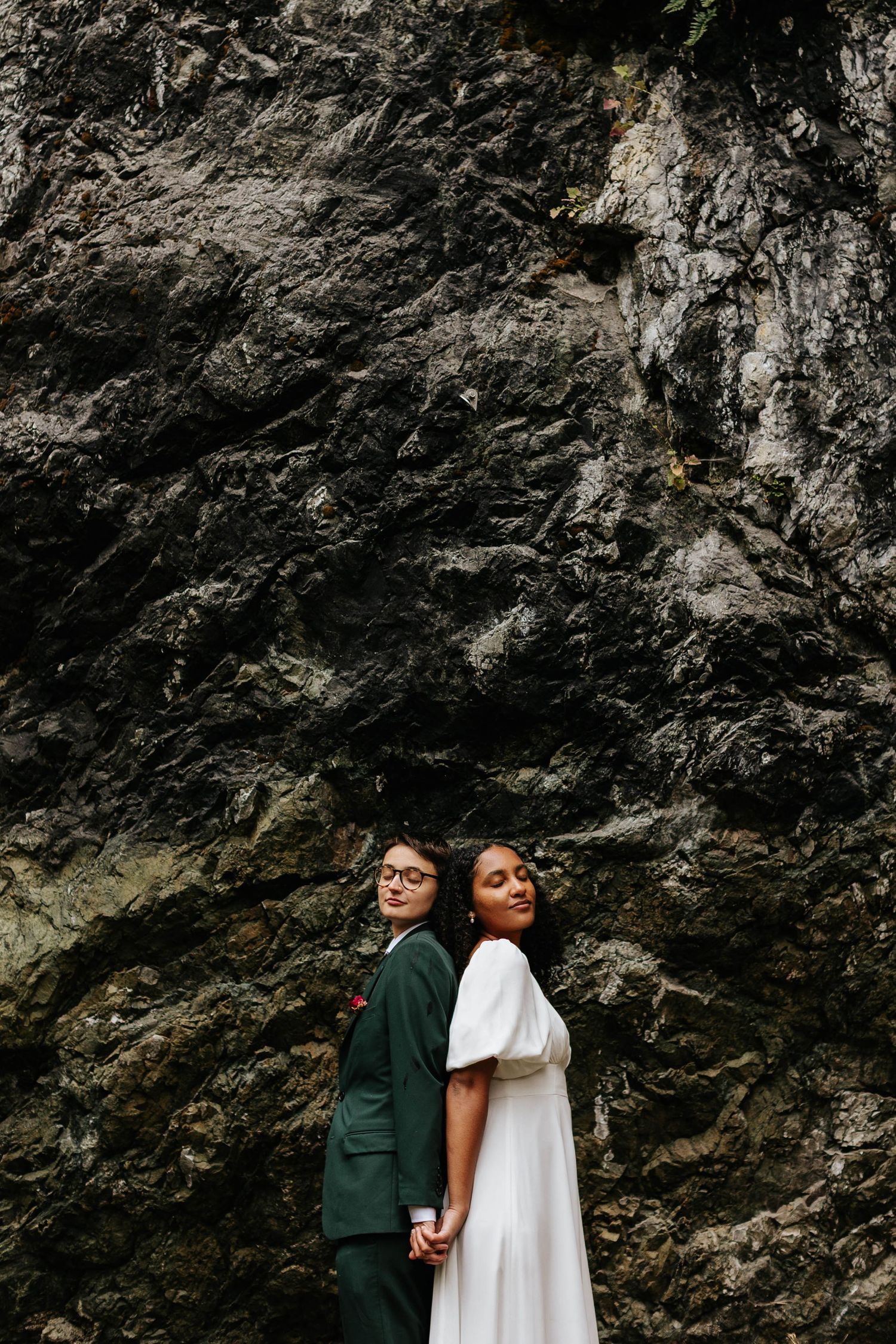 Bride in white dress and groom in green suit stand back-to-back against a dramatic dark rock wall.