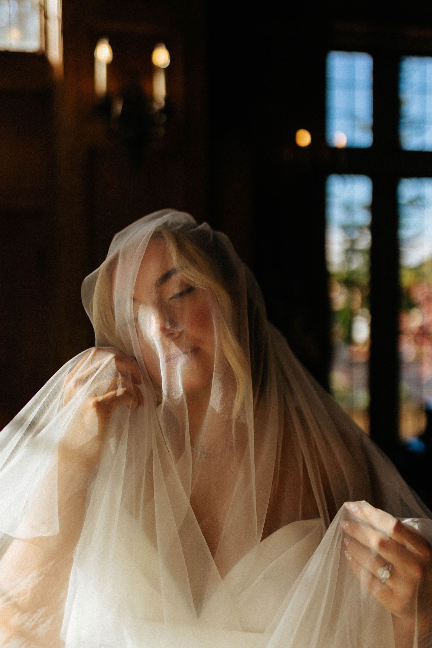 Blonde bride draped in a sheer veil poses in soft candlelit indoor light with eyes gently closed.