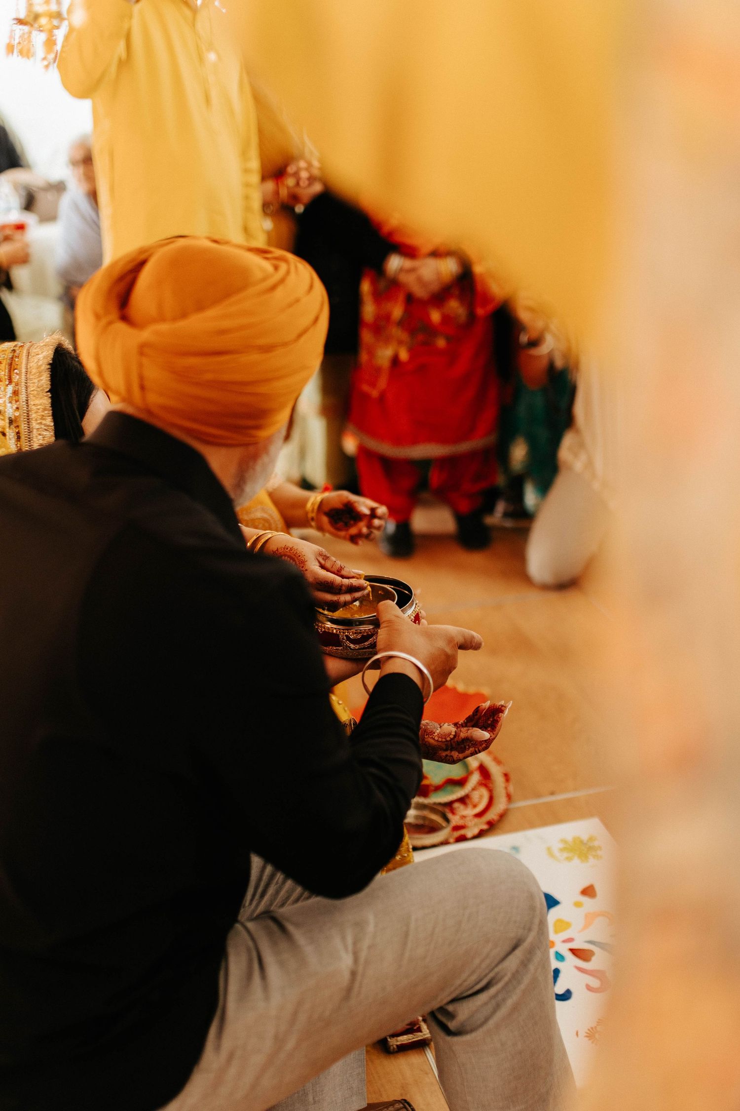 Person wearing orange turban seated during traditional Sikh ceremony with decorative items nearby.