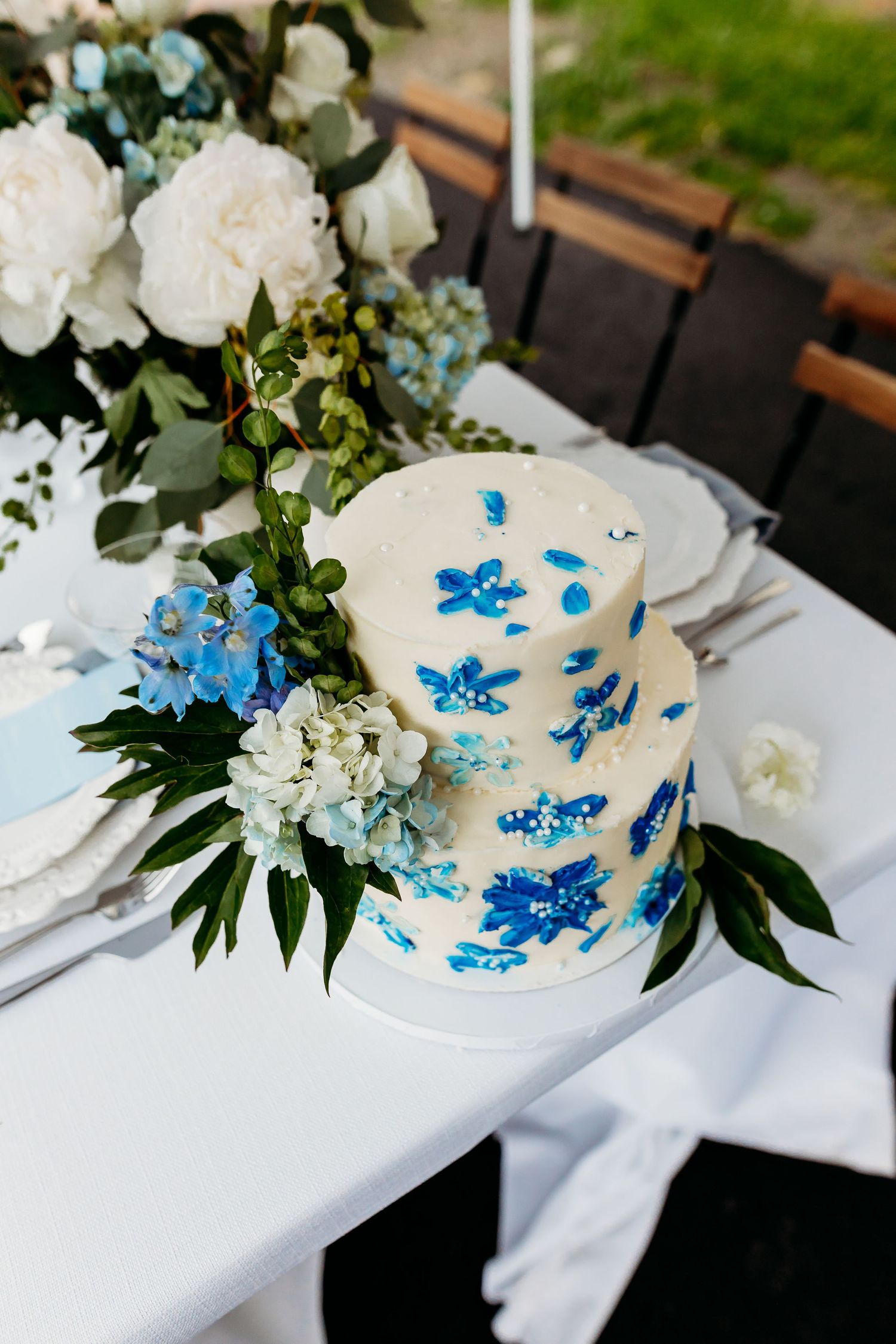 White wedding cake decorated with delicate blue floral designs and fresh flowers on a table setting with white linens.