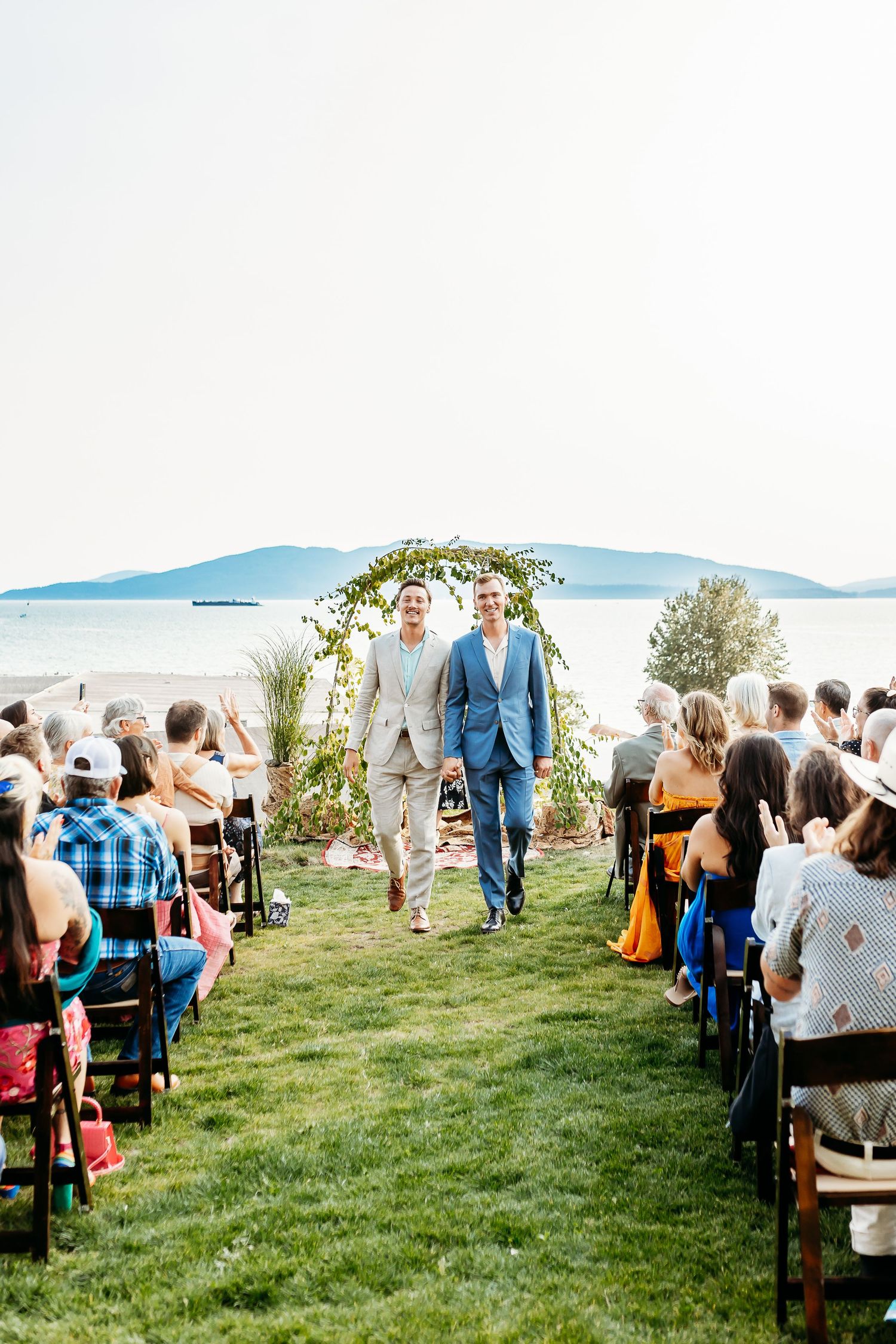 Beautiful outdoor wedding ceremony on a grassy lawn overlooking blue mountains and water, with guests seated on both sides of the aisle.