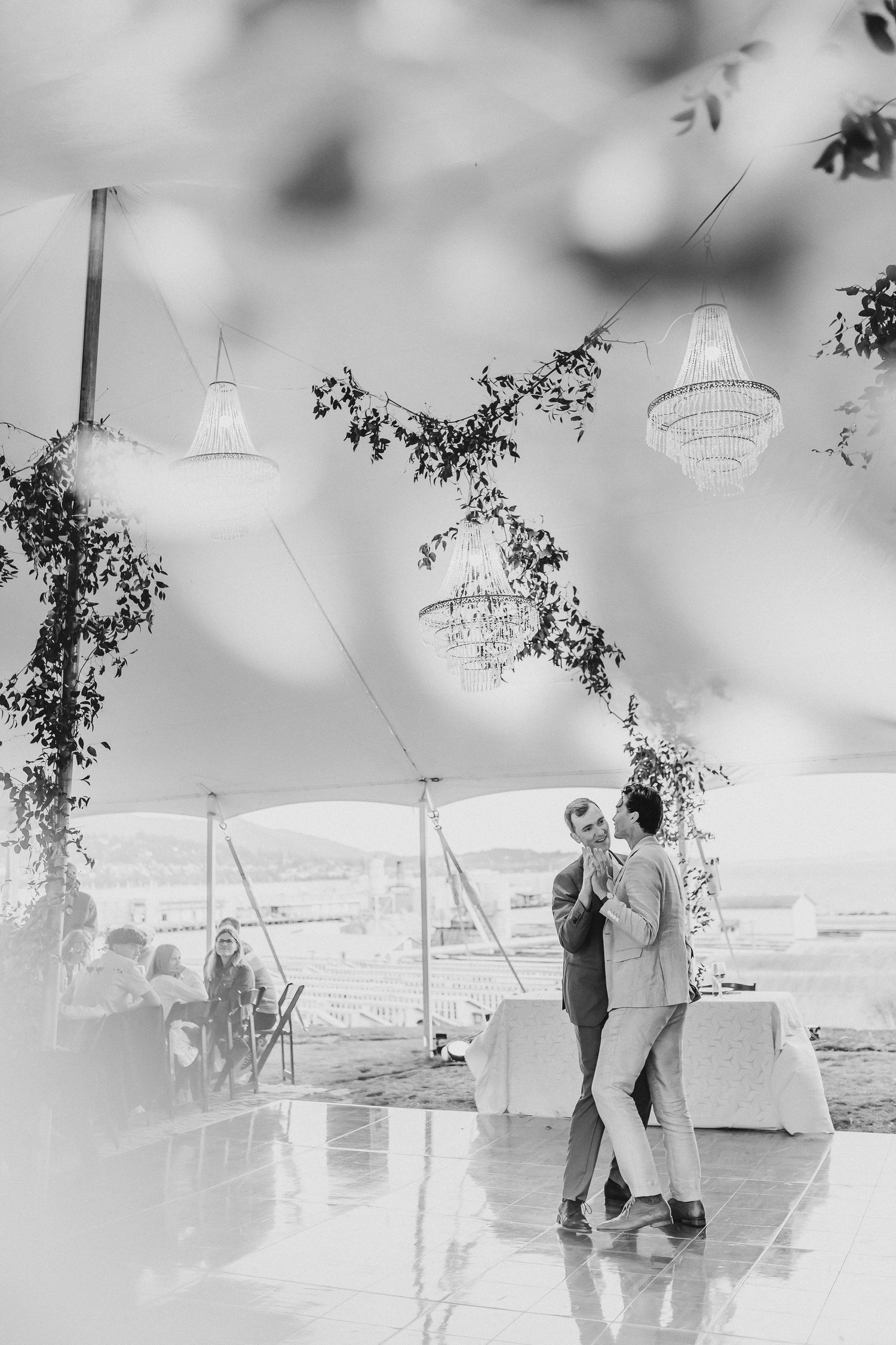 A couple dances beneath crystal chandeliers in an elegant tent at an outdoor wedding reception with greenery garlands.