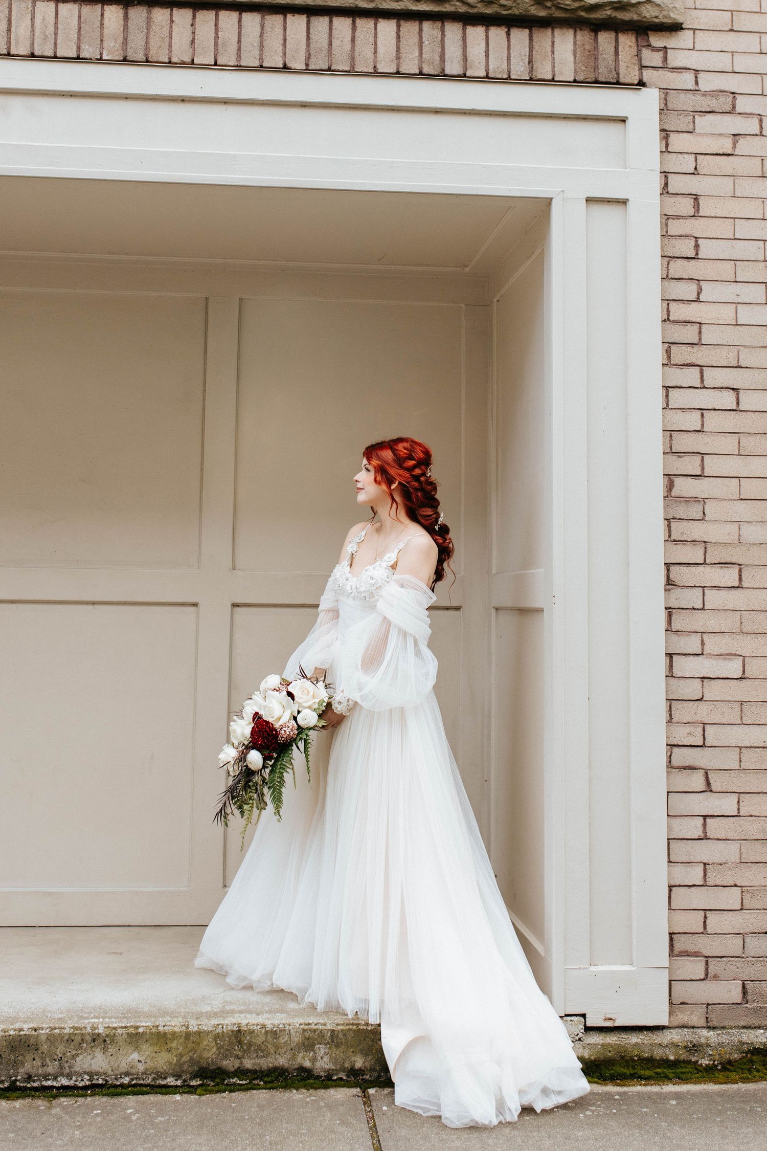 Red-haired bride in an off-shoulder white tulle gown holding a bouquet stands in a brick building doorway.