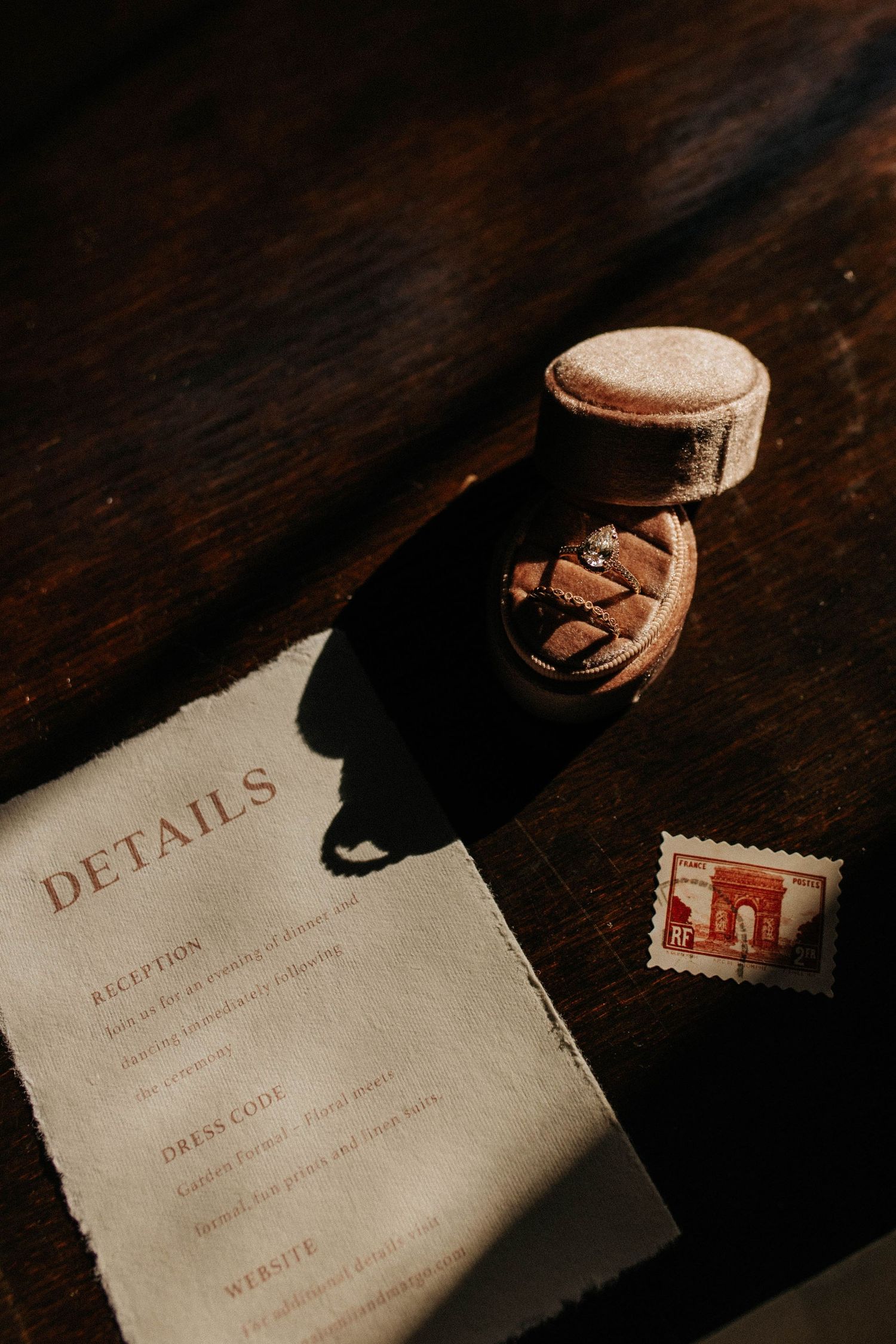Wedding details flat lay with a velvet ring box, diamond ring, vintage postage stamp, and details card on dark wood.