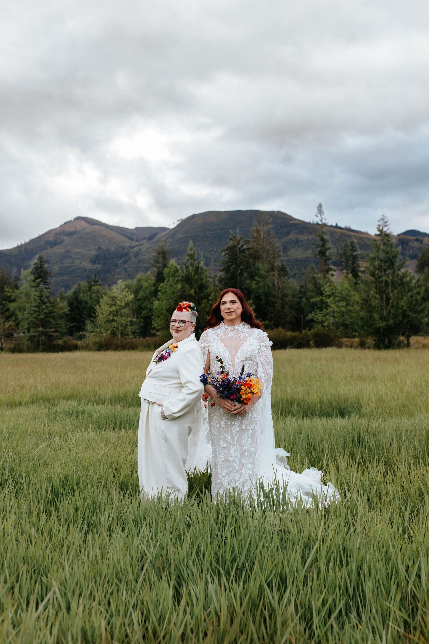 Two brides pose in white dresses holding bouquets in a grassy field with mountains in the background.