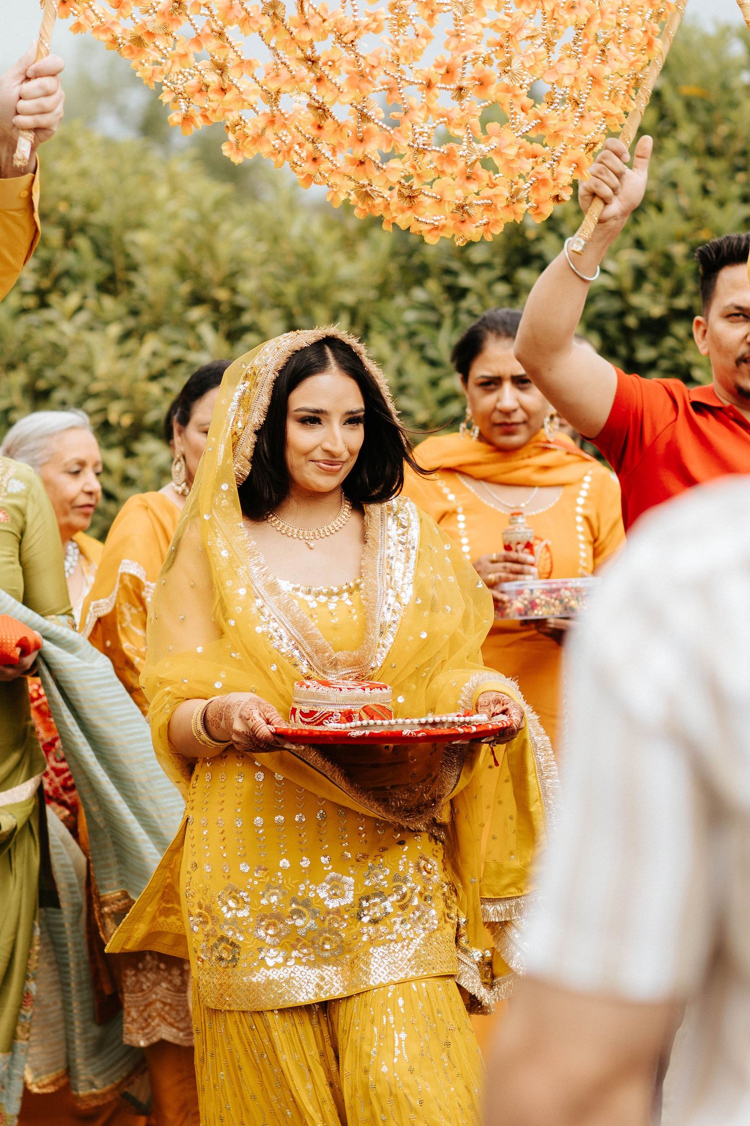 Guests in vibrant yellow traditional Indian attire celebrate at an outdoor wedding ceremony with orange decorative elements.