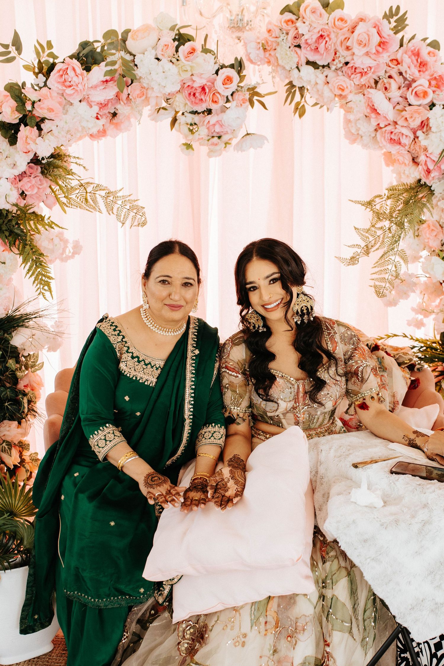 Two people sit together under a pink floral arch wearing elegant traditional South Asian attire.