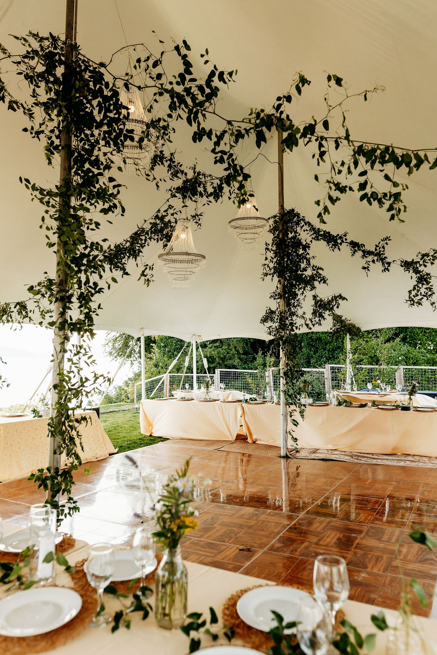 Elegant wedding tent interior decorated with greenery vines, wooden dance floor, and white table settings for reception.
