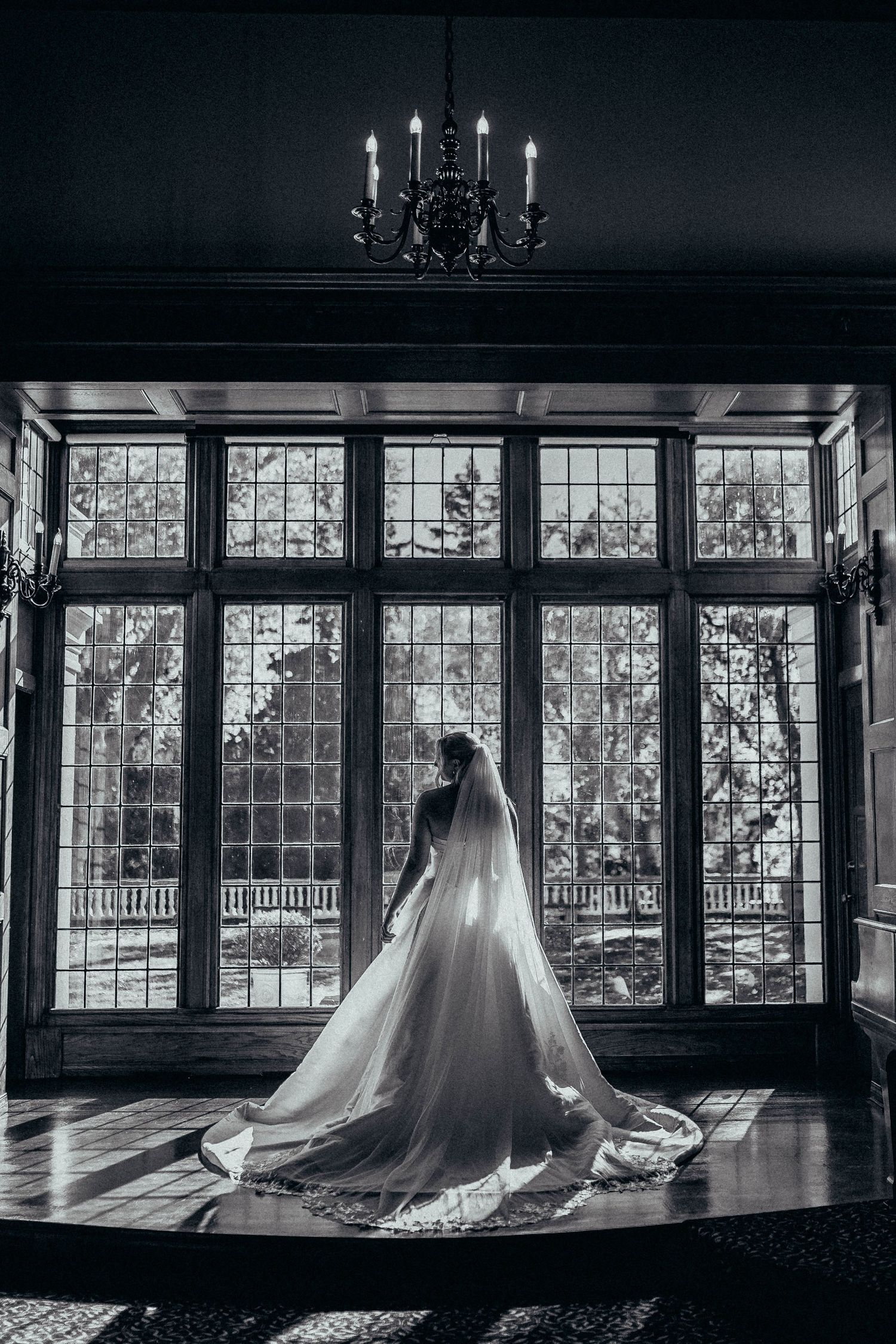 Dramatic black and white silhouette of bride in cathedral veil standing before large leaded glass windows.