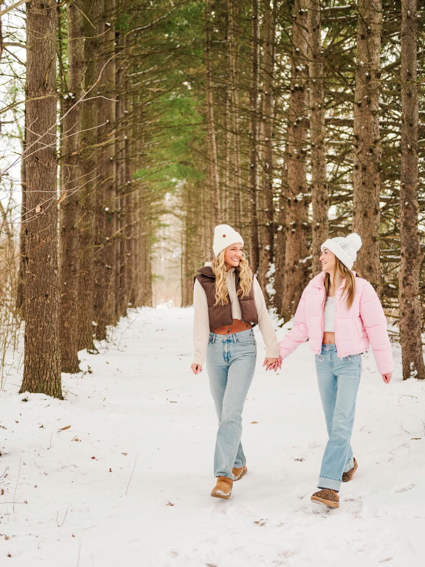 Two women in winter outfits walk hand-in-hand through a snowy pine tree path wearing beanies and puffer jackets.