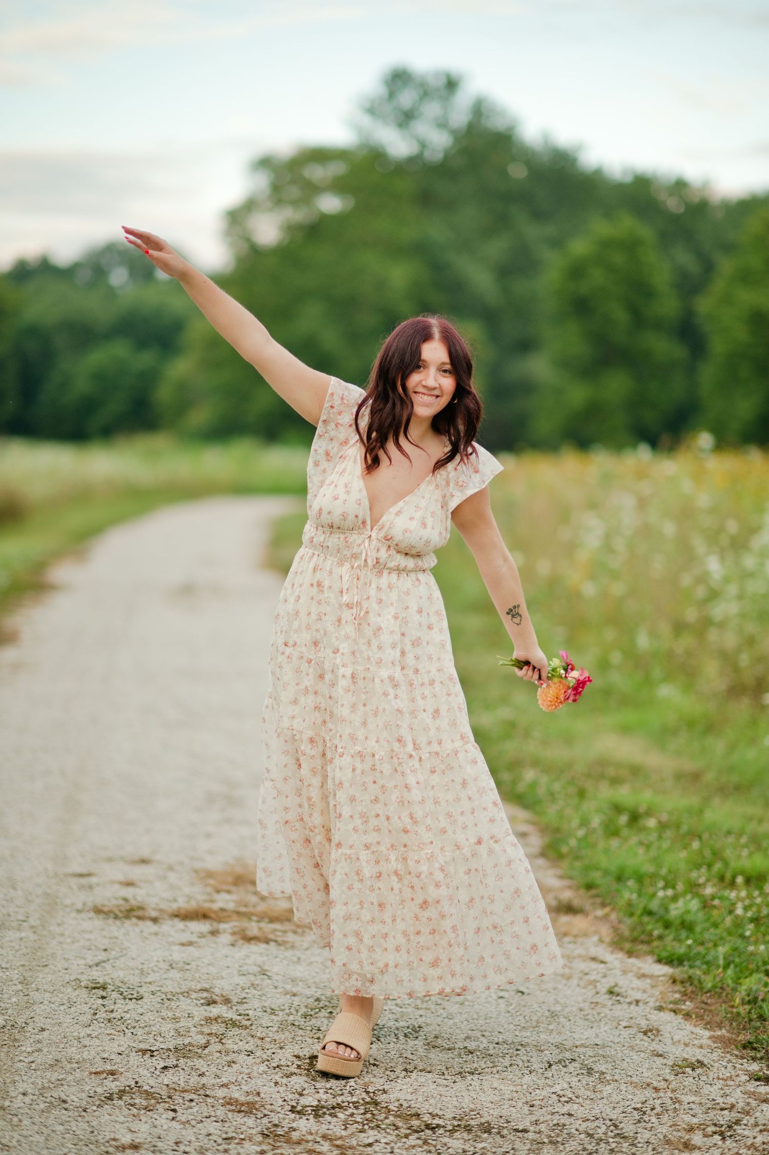 Happy brunette woman in floral dress twirling on a rural gravel path with flowers.