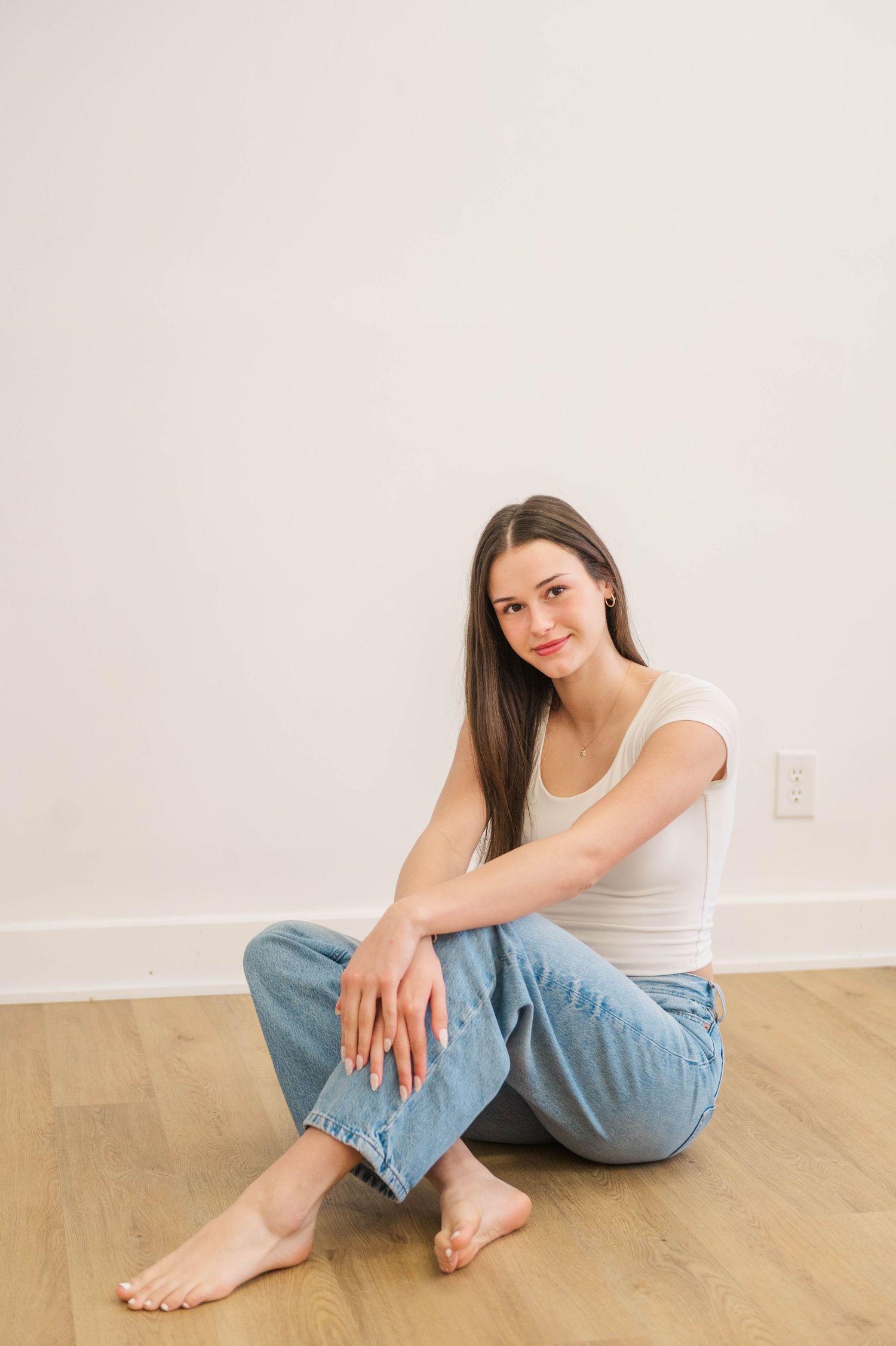 Young woman in white tank top and blue jeans sitting barefoot on hardwood floor against white wall.