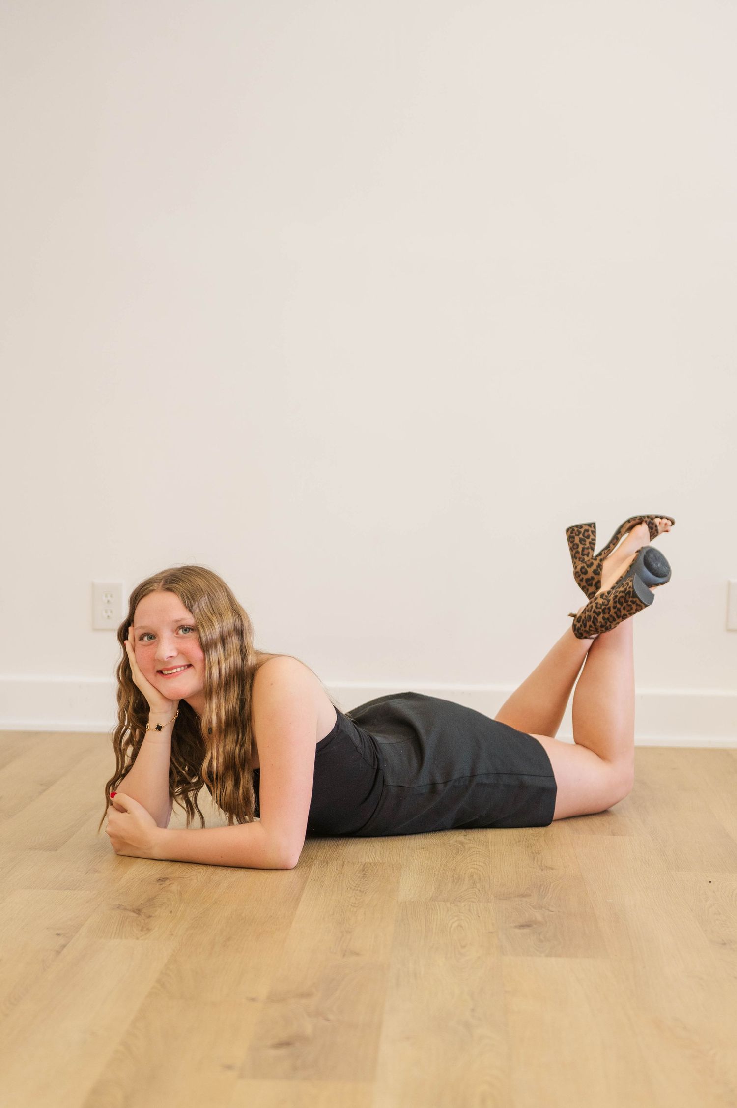 Young woman in black mini dress and leopard heels lying on hardwood floor smiling with legs crossed up.