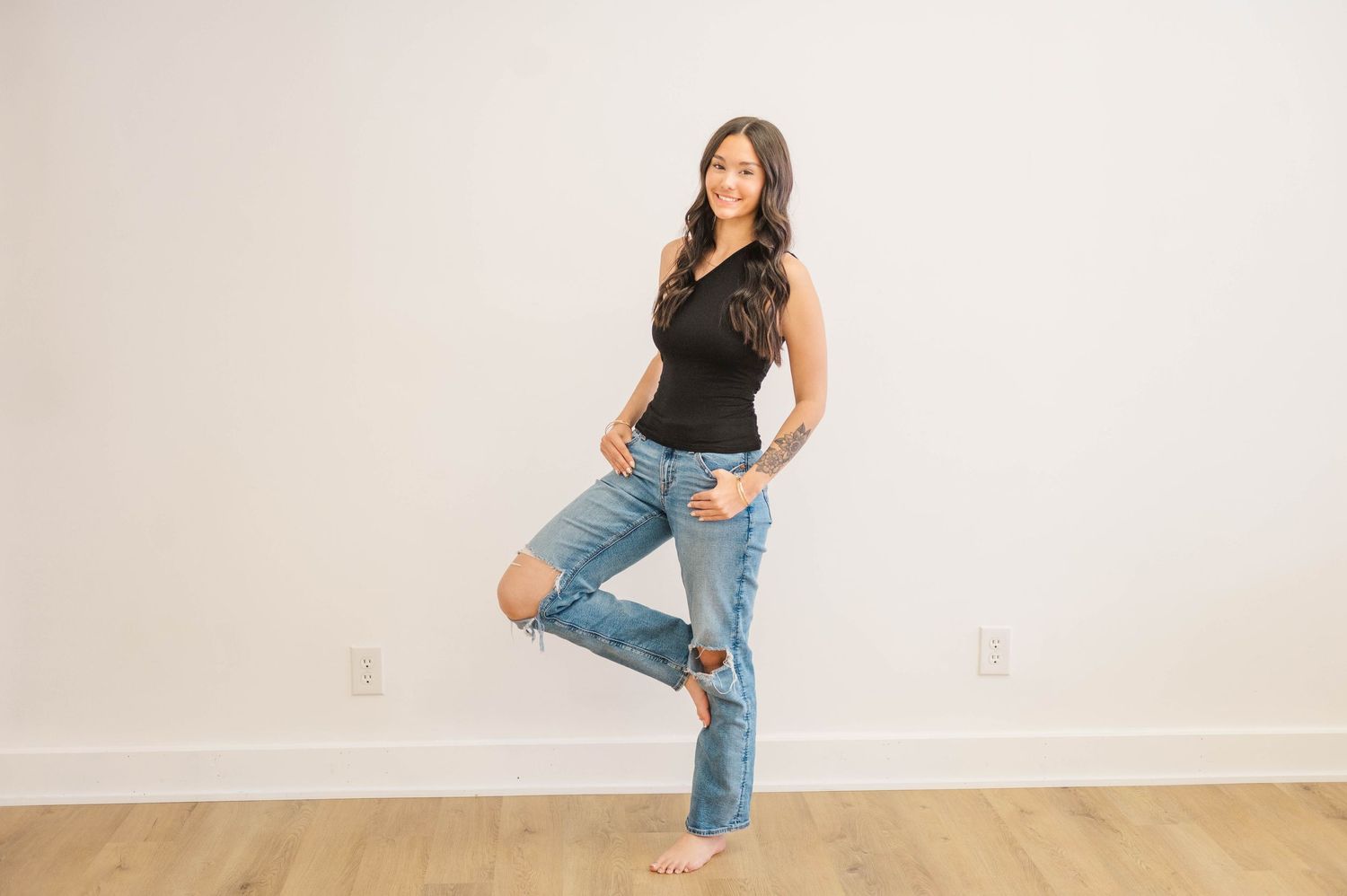 Woman in black one-shoulder top and ripped jeans posing on one leg against white wall.