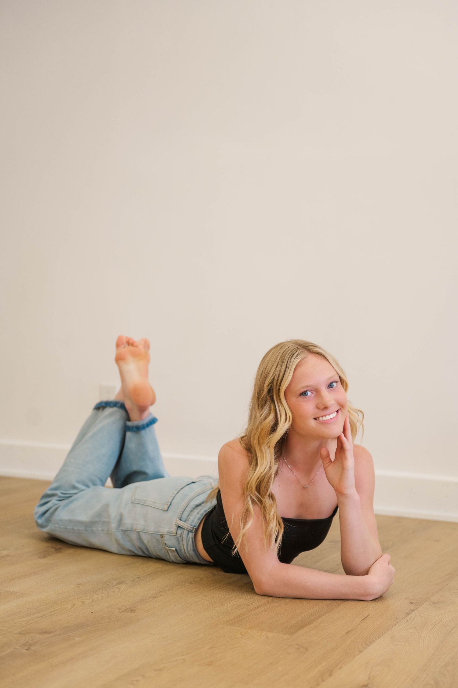 Blonde woman in black top and light jeans lying on hardwood floor looking up smiling.