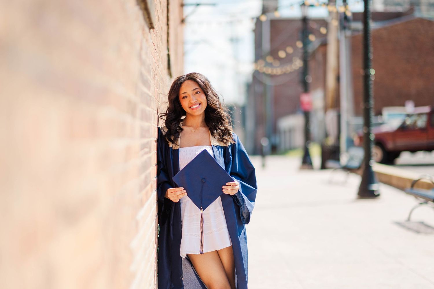Young woman in navy graduation gown holding cap while leaning against brick wall outdoors.