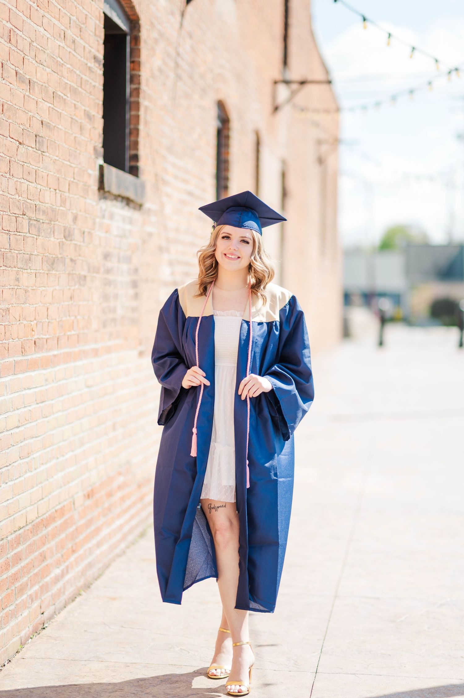 Blonde graduate in navy cap and gown smiling while walking near brick wall on sunny day.