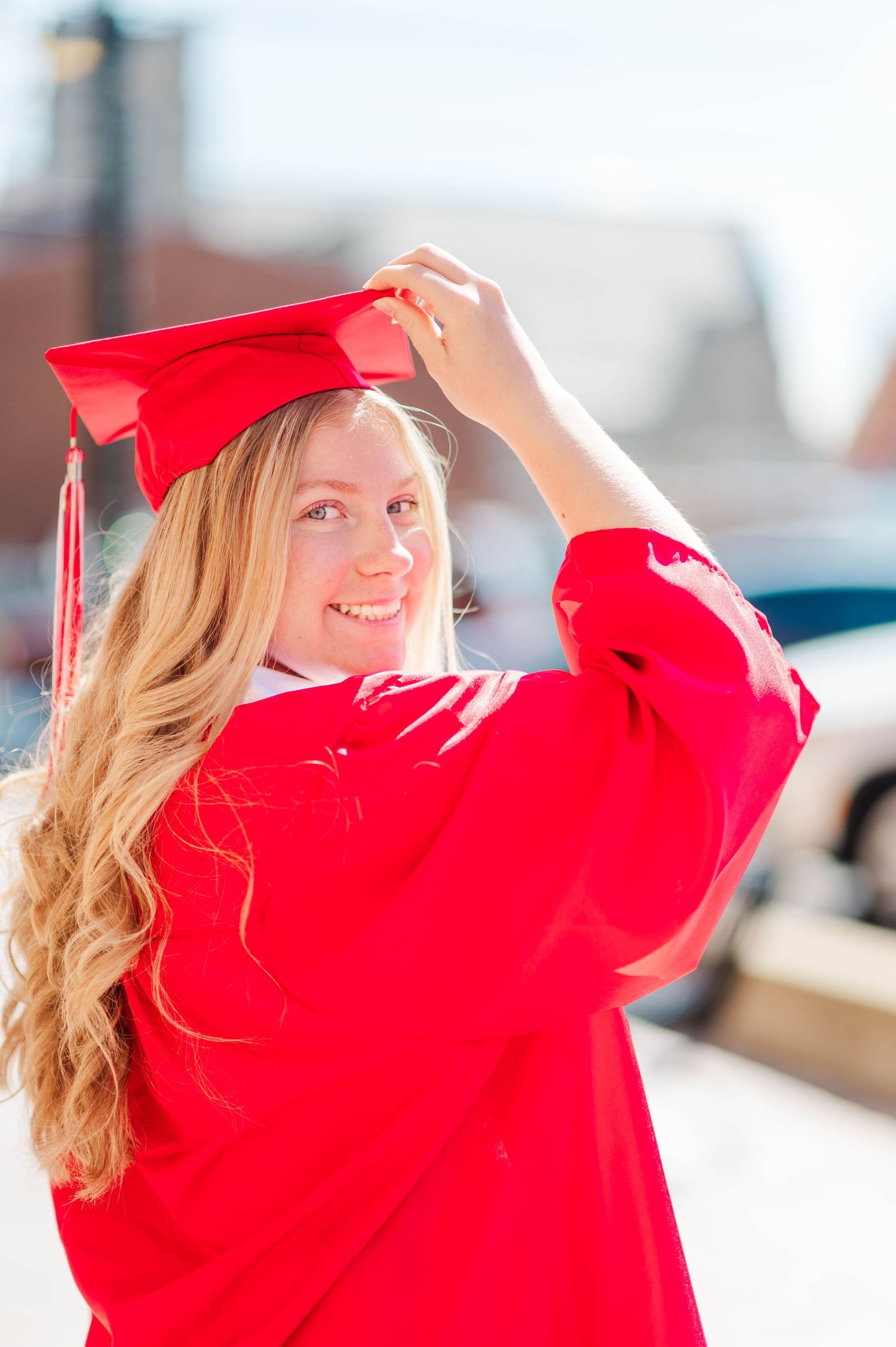 Smiling blonde woman in red graduation cap and gown posing outdoors on a sunny day.