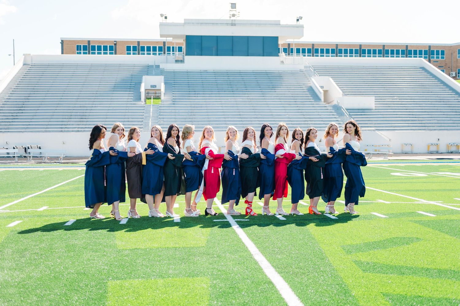 Smiling graduates in navy gowns holding diplomas on a sunny football field stadium.