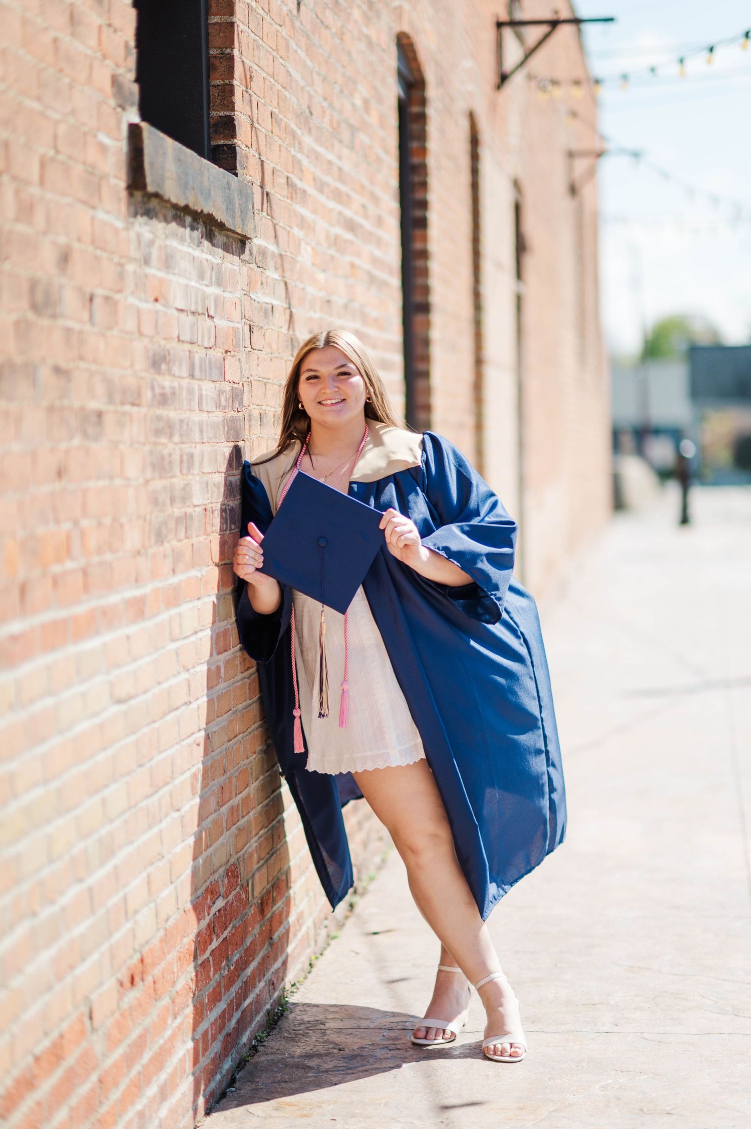 Happy female graduate leans against brick wall holding navy cap and gown in sunny outdoor alley.