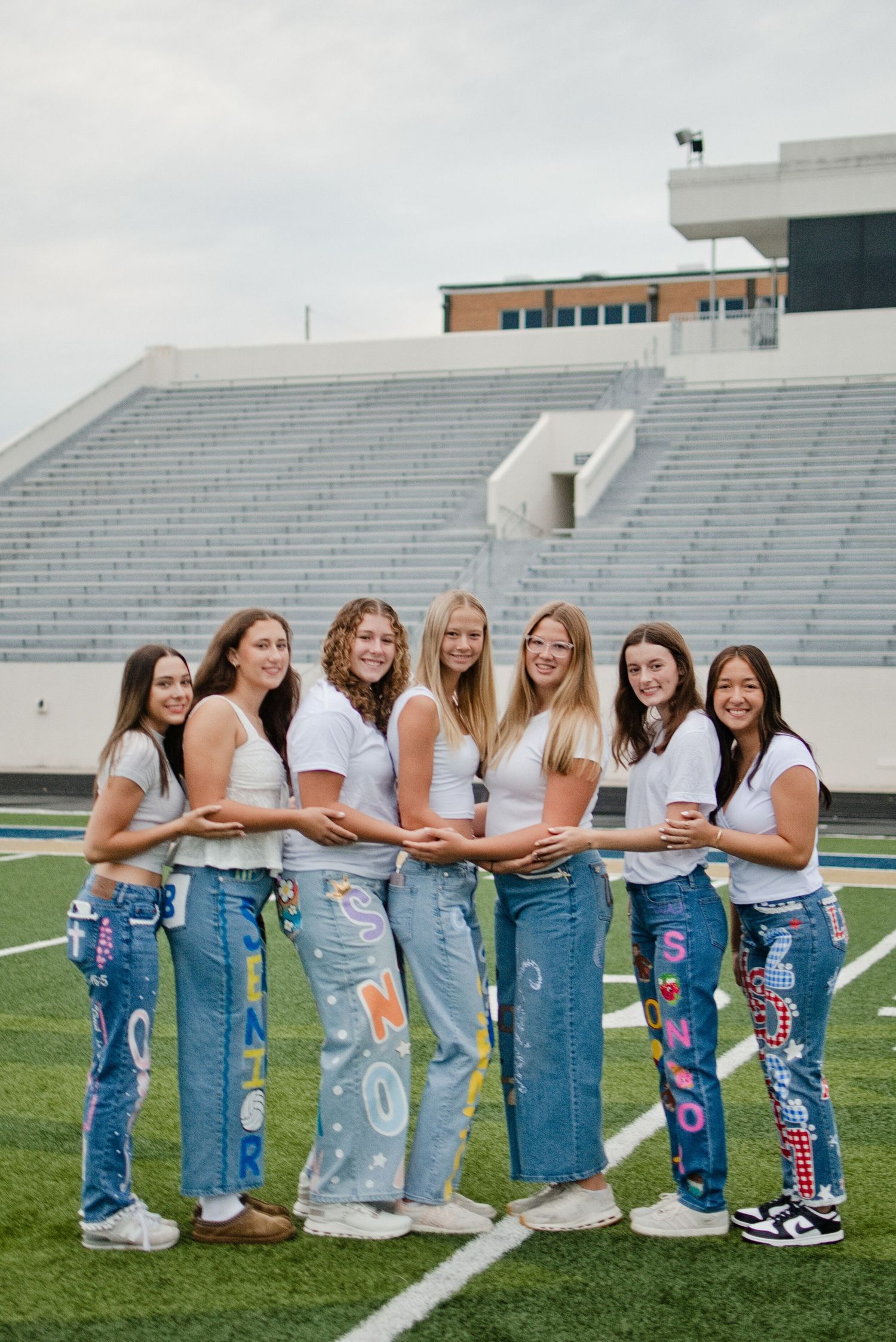 Seven girls in white tees and custom painted senior jeans pose together on a football field stadium.