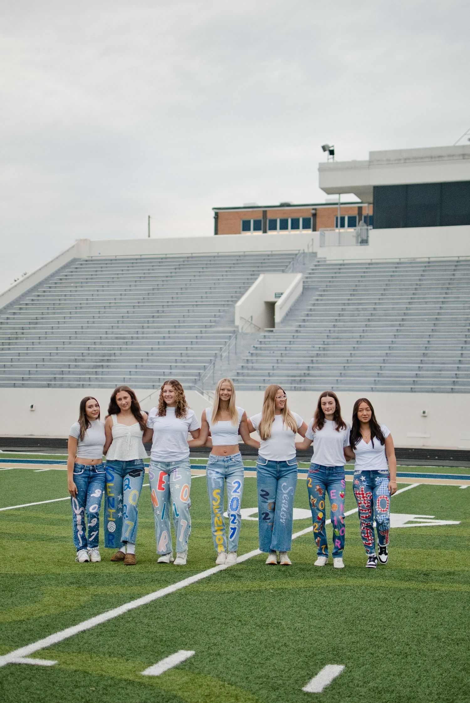 Seven girls in white tops and decorated jeans pose together on a football field in front of stadium bleachers.