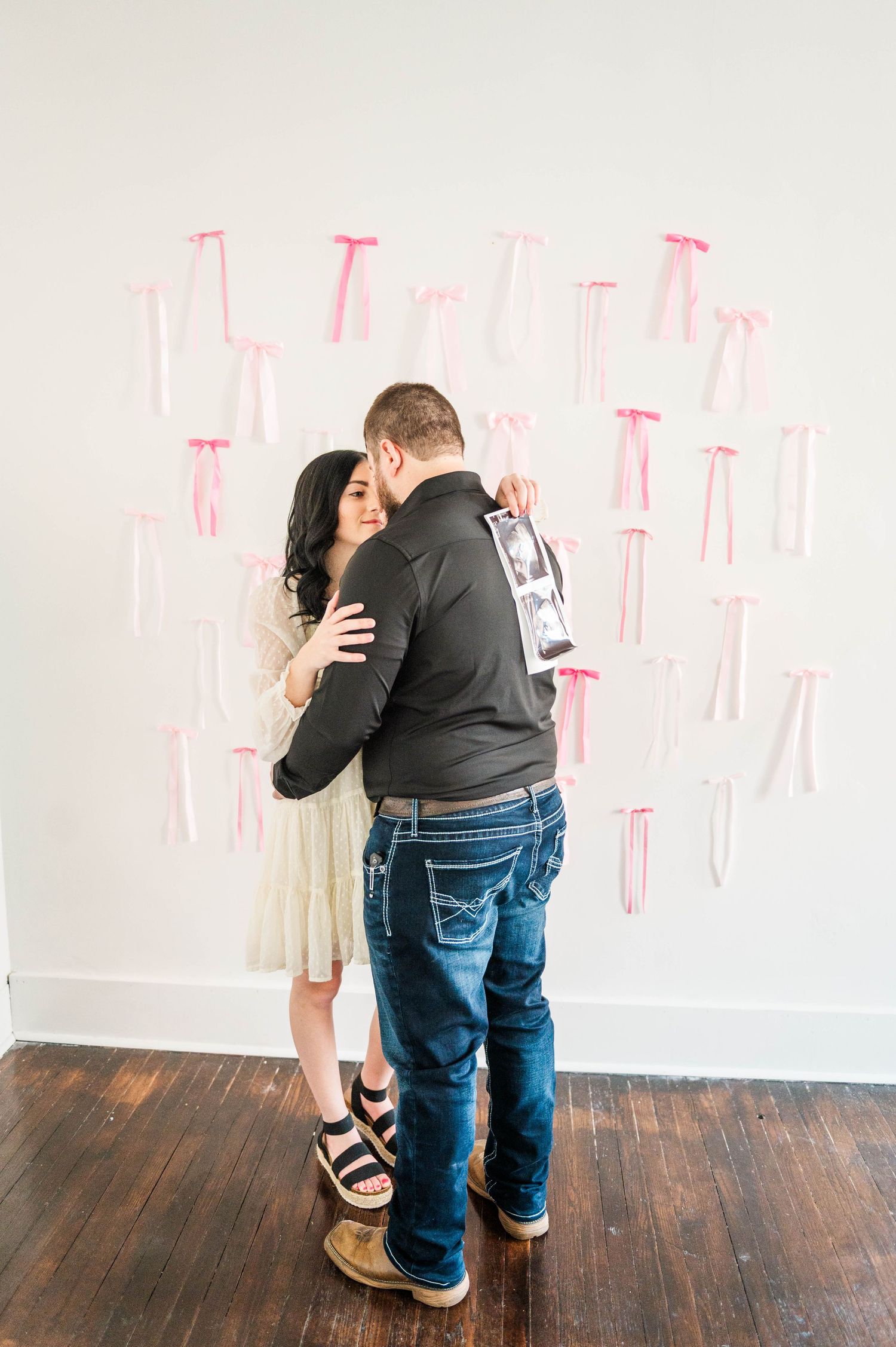 A couple embraces while holding ultrasound photos against a white wall with pink decorations.