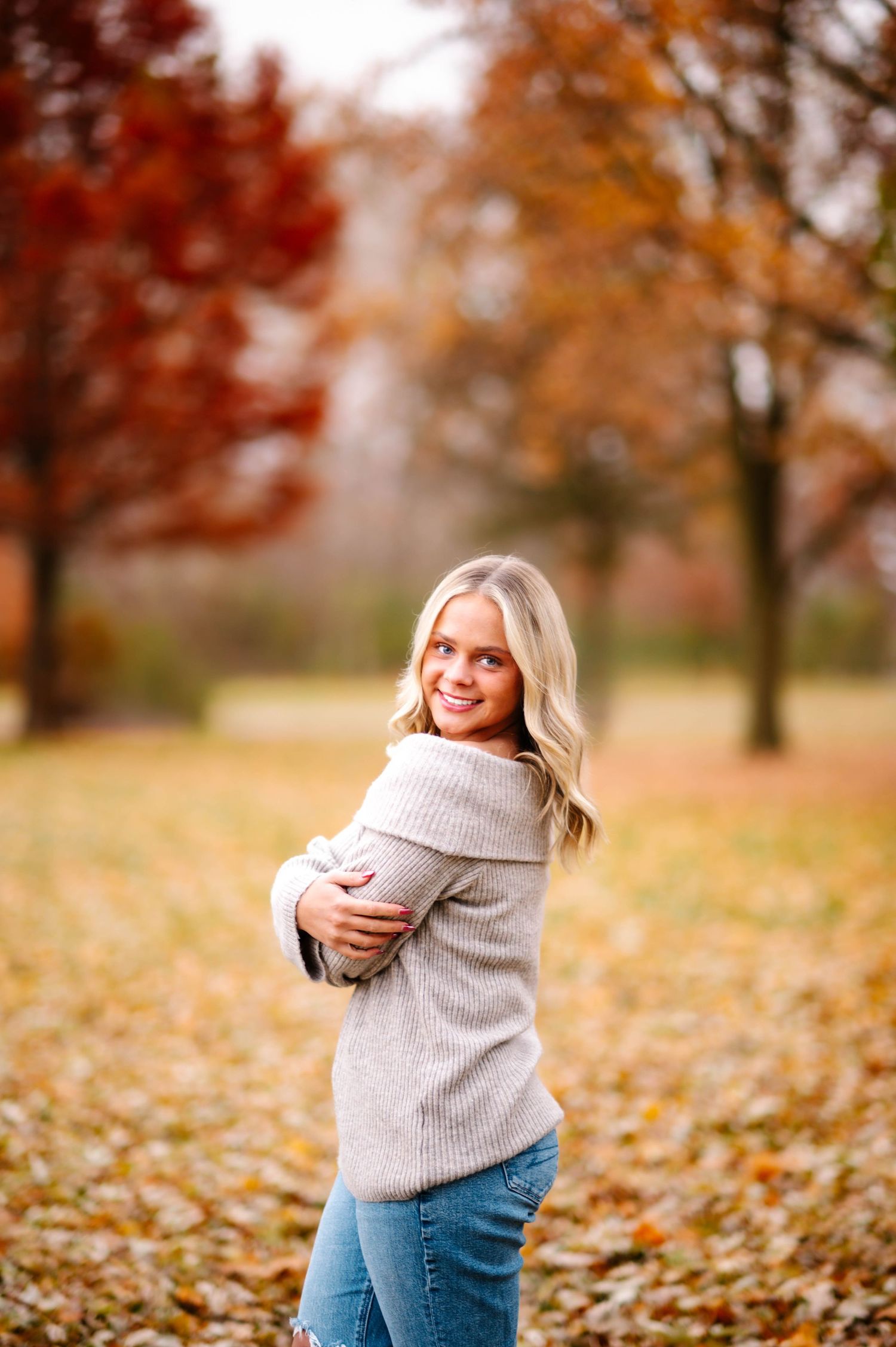 Person in cozy gray sweater and jeans poses playfully in autumn park with fallen leaves on ground.