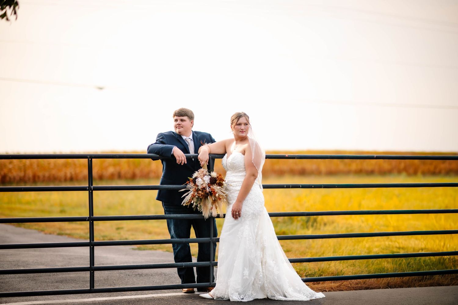 Wedding couple poses together against rustic fence with scenic countryside view.