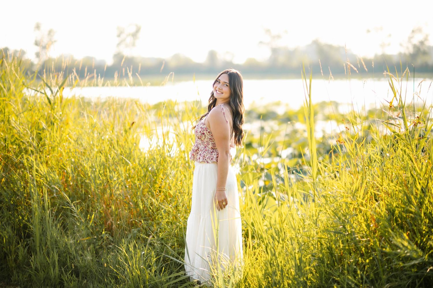 Young woman in white maxi skirt and floral top standing in golden sunlit meadow near a lake.