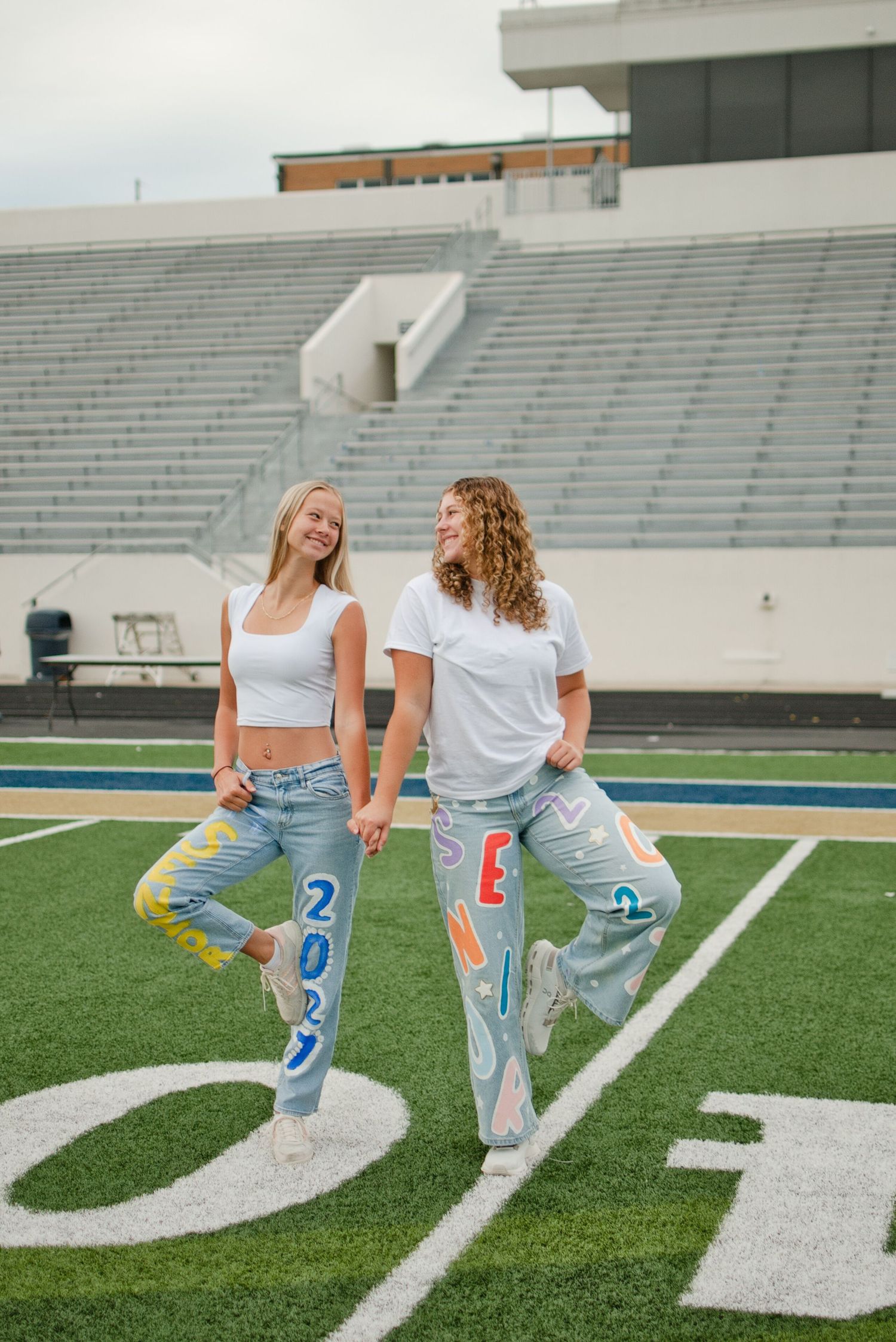 Two girls in white tops and painted senior 2027 jeans laugh while posing on a football field.