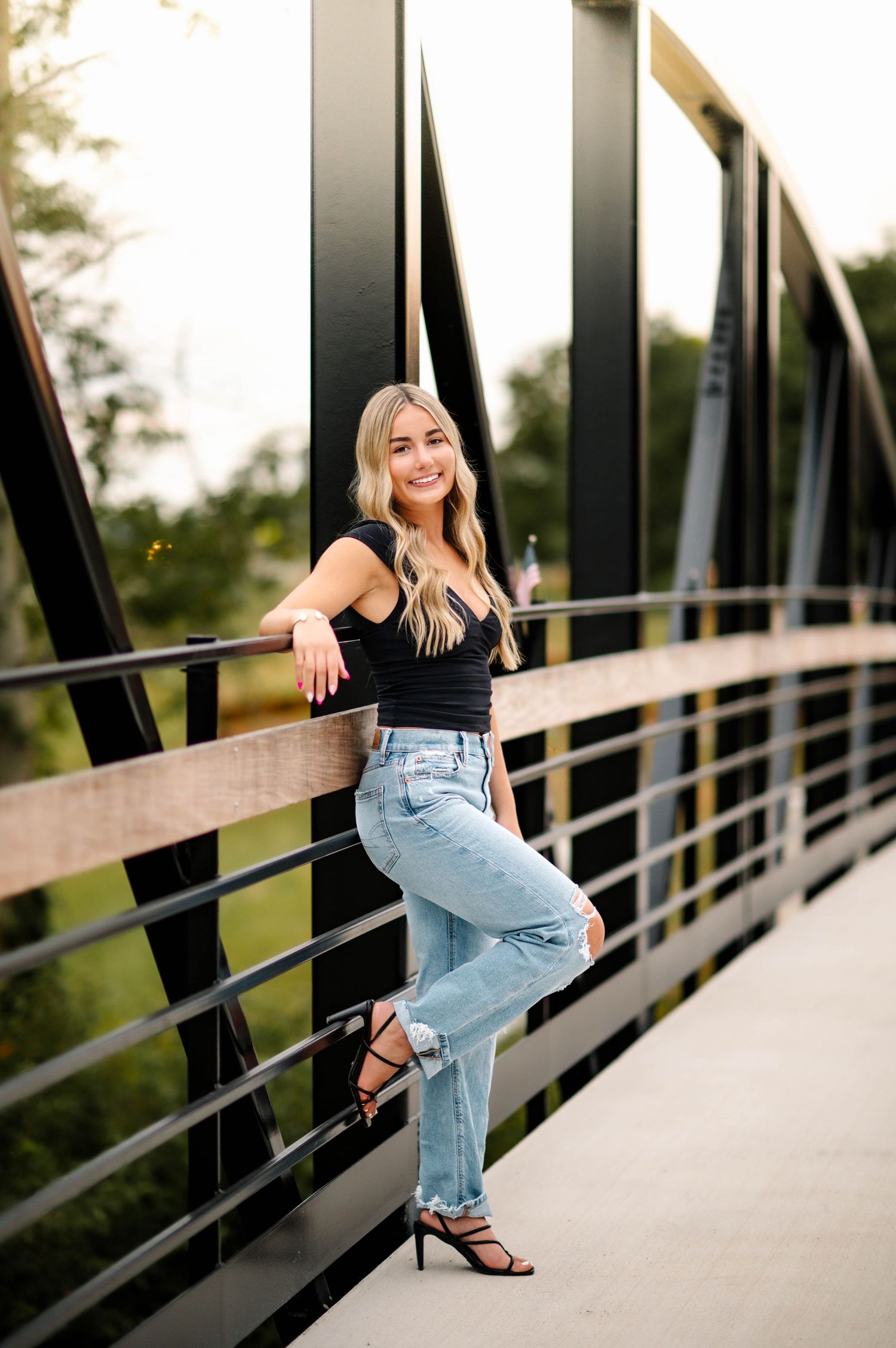 Stylish casual outfit featuring black top and jeans while posing on metal bridge railing.