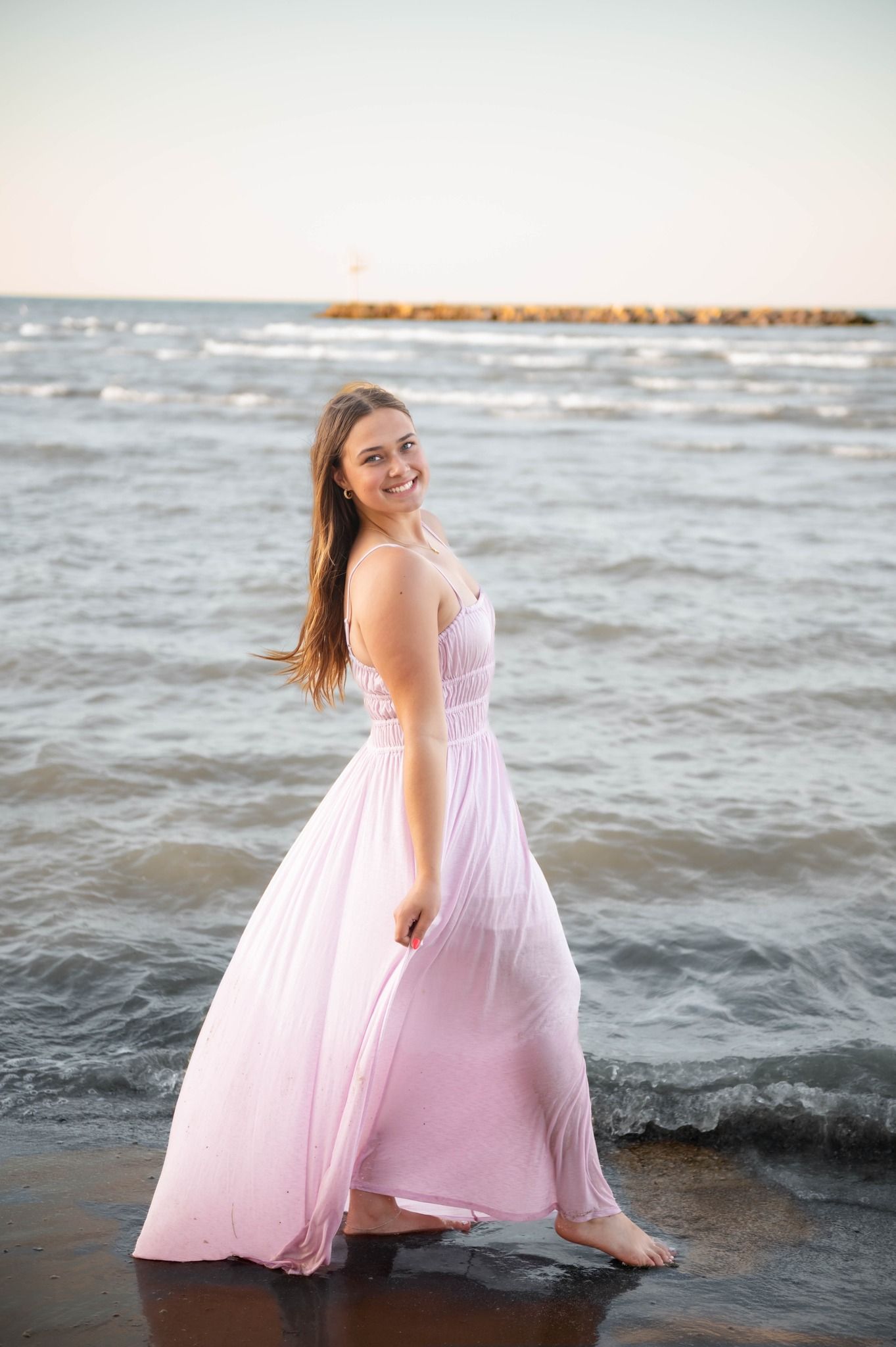 A person in a flowing pink dress poses playfully on a beach at sunset with waves in the background.