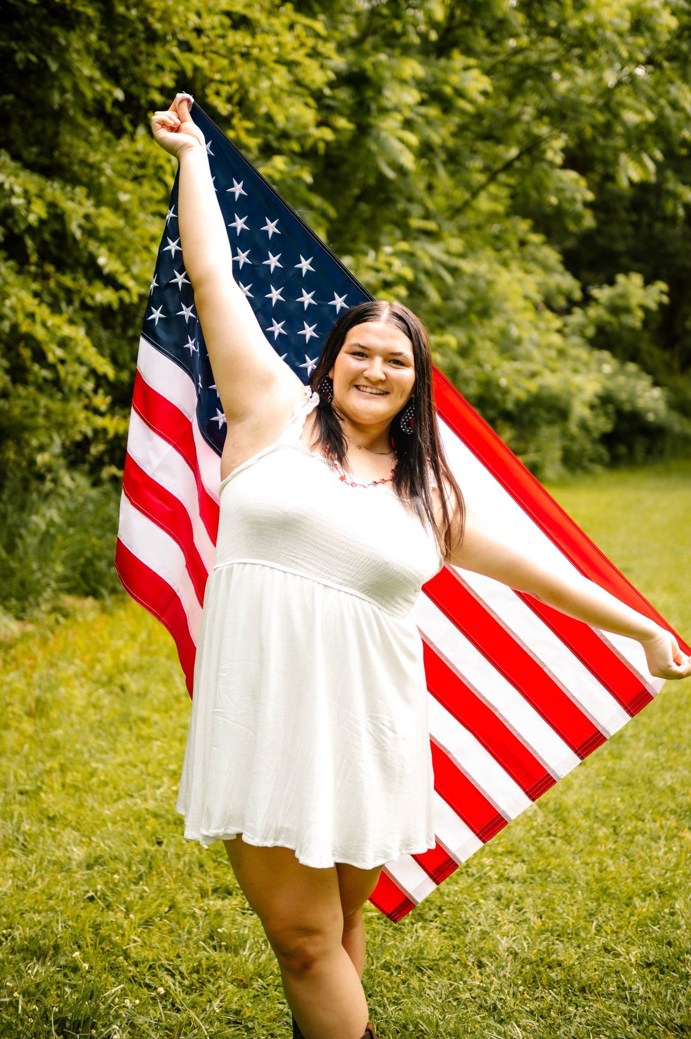 Woman in white dress joyfully poses with American flag in outdoor summer setting.