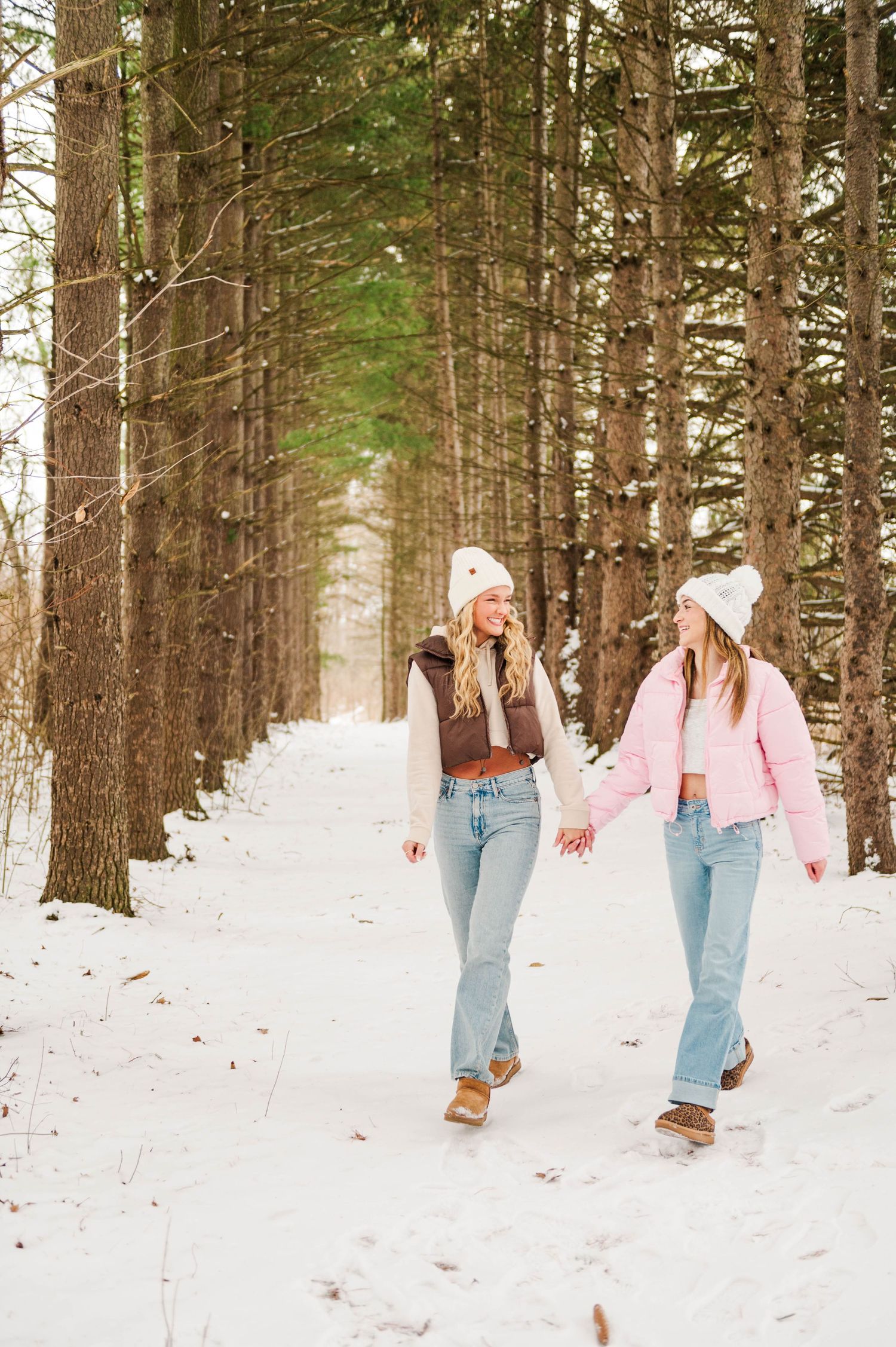 Two friends in winter jackets and beanies walk together through a snow-covered forest path.