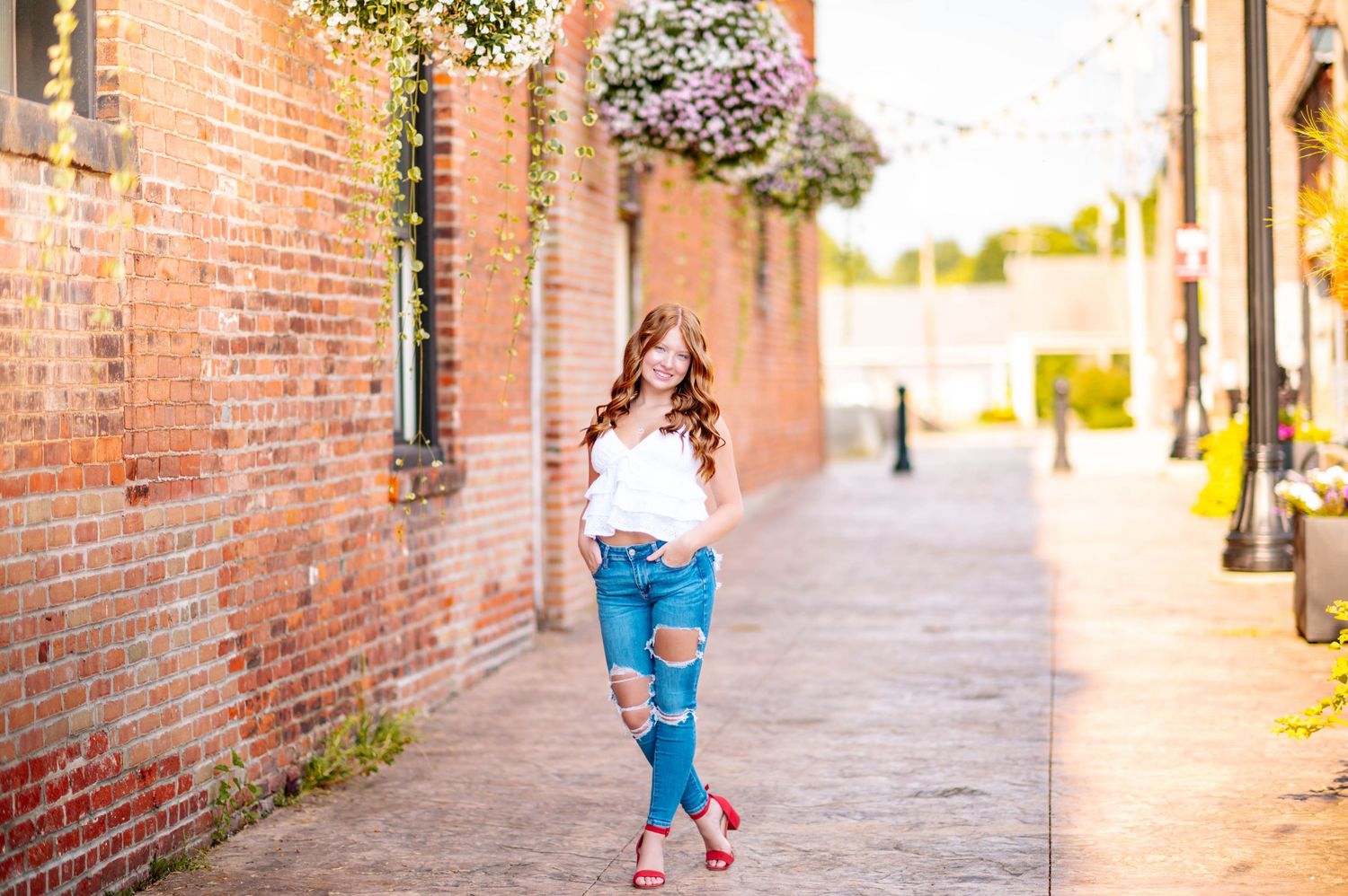 Young woman in ripped jeans and white top poses on a charming brick alley with hanging flower baskets.