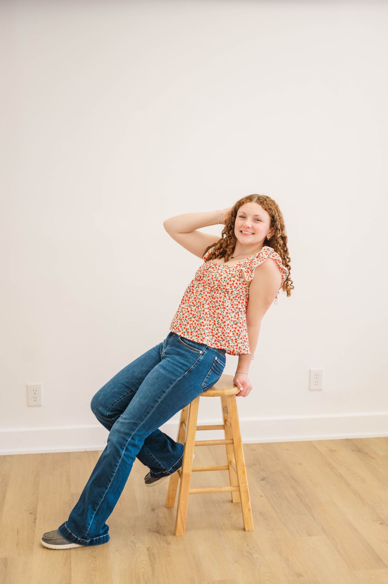 Young girl in floral top and flare jeans sitting on wooden stool with hand in hair.