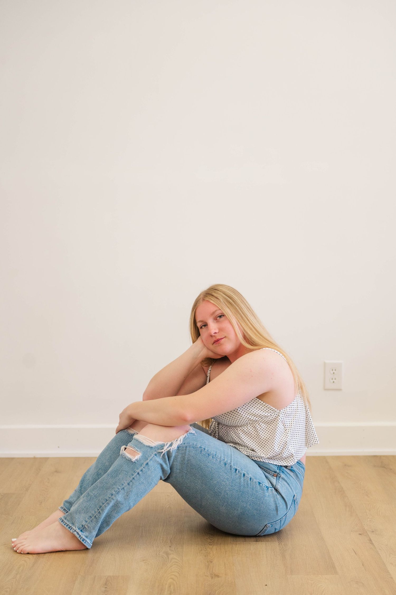 Young blonde woman sitting on hardwood floor in ripped jeans and striped tank top against white wall.