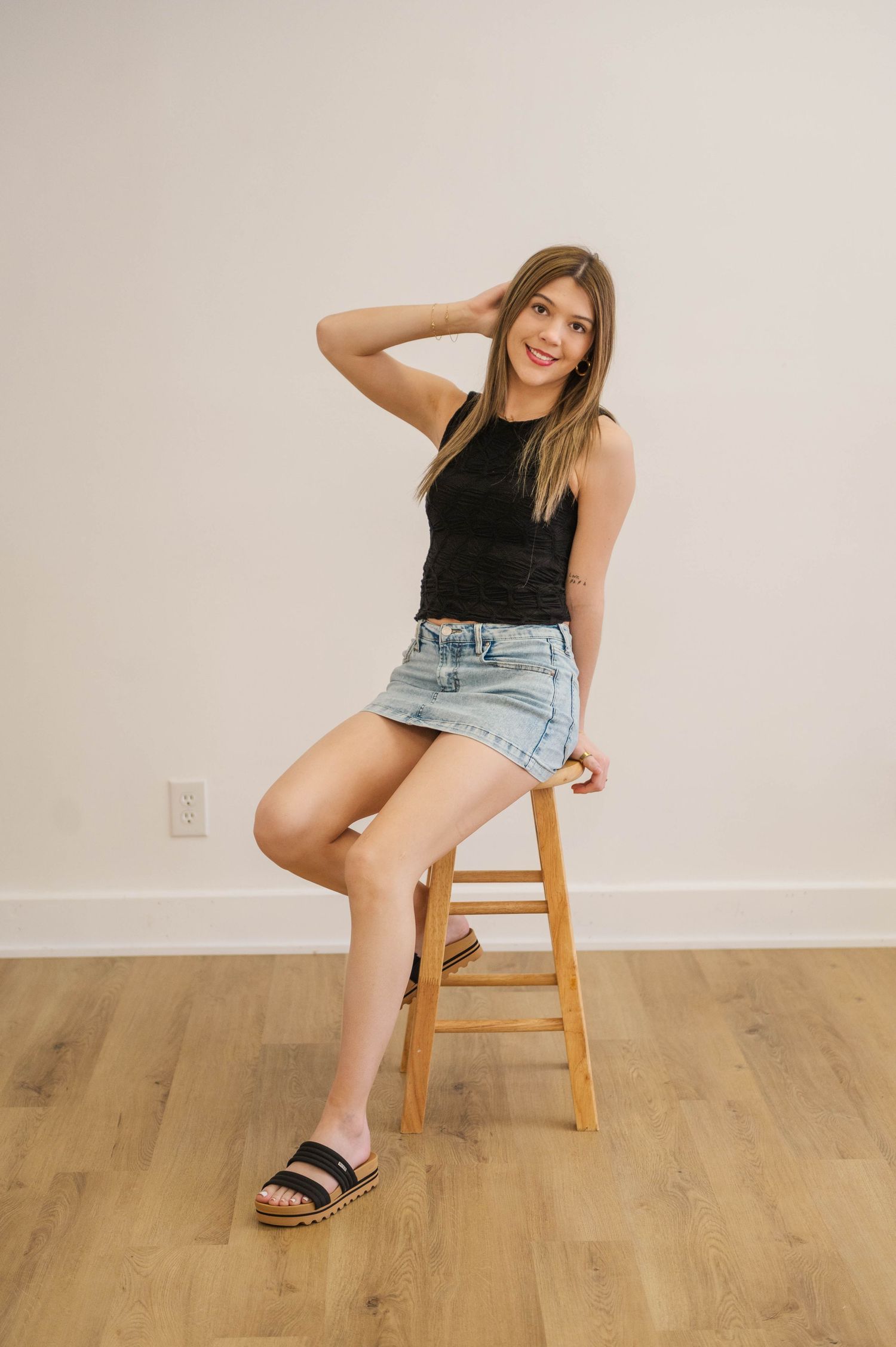 Young woman in black tank top and denim mini skirt sitting on wooden stool.