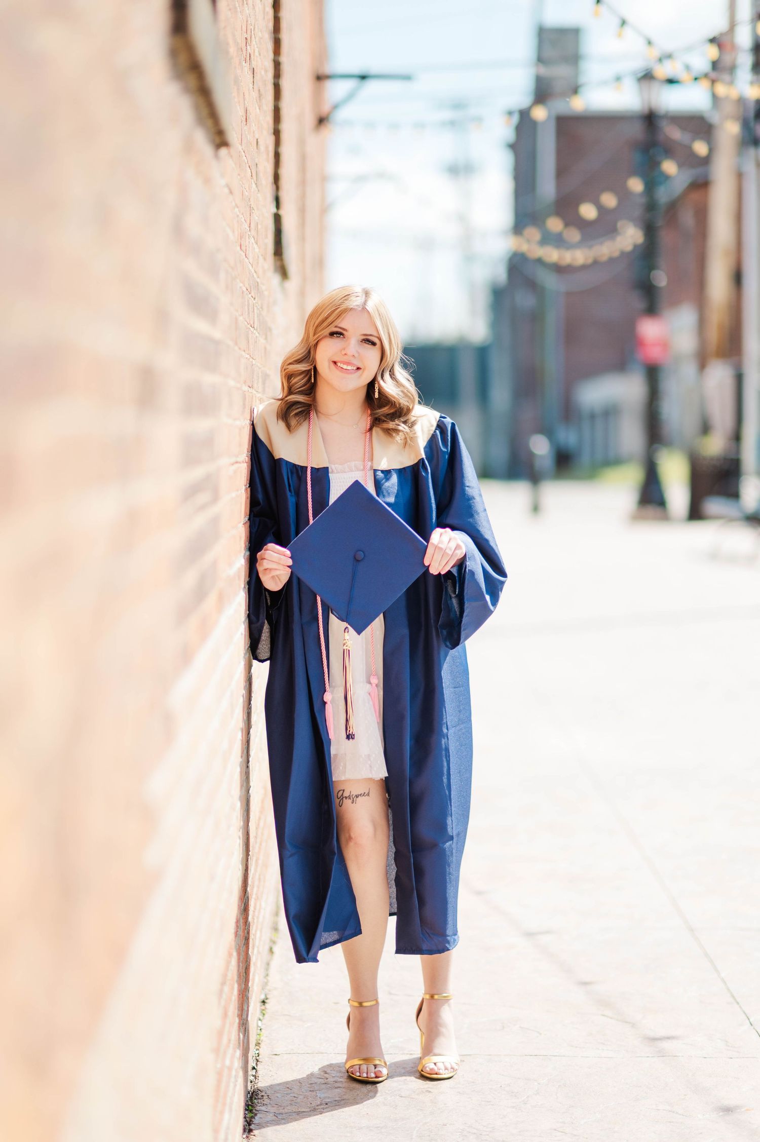 Smiling graduate in blue cap and gown leaning against brick wall outdoors on sunny day.
