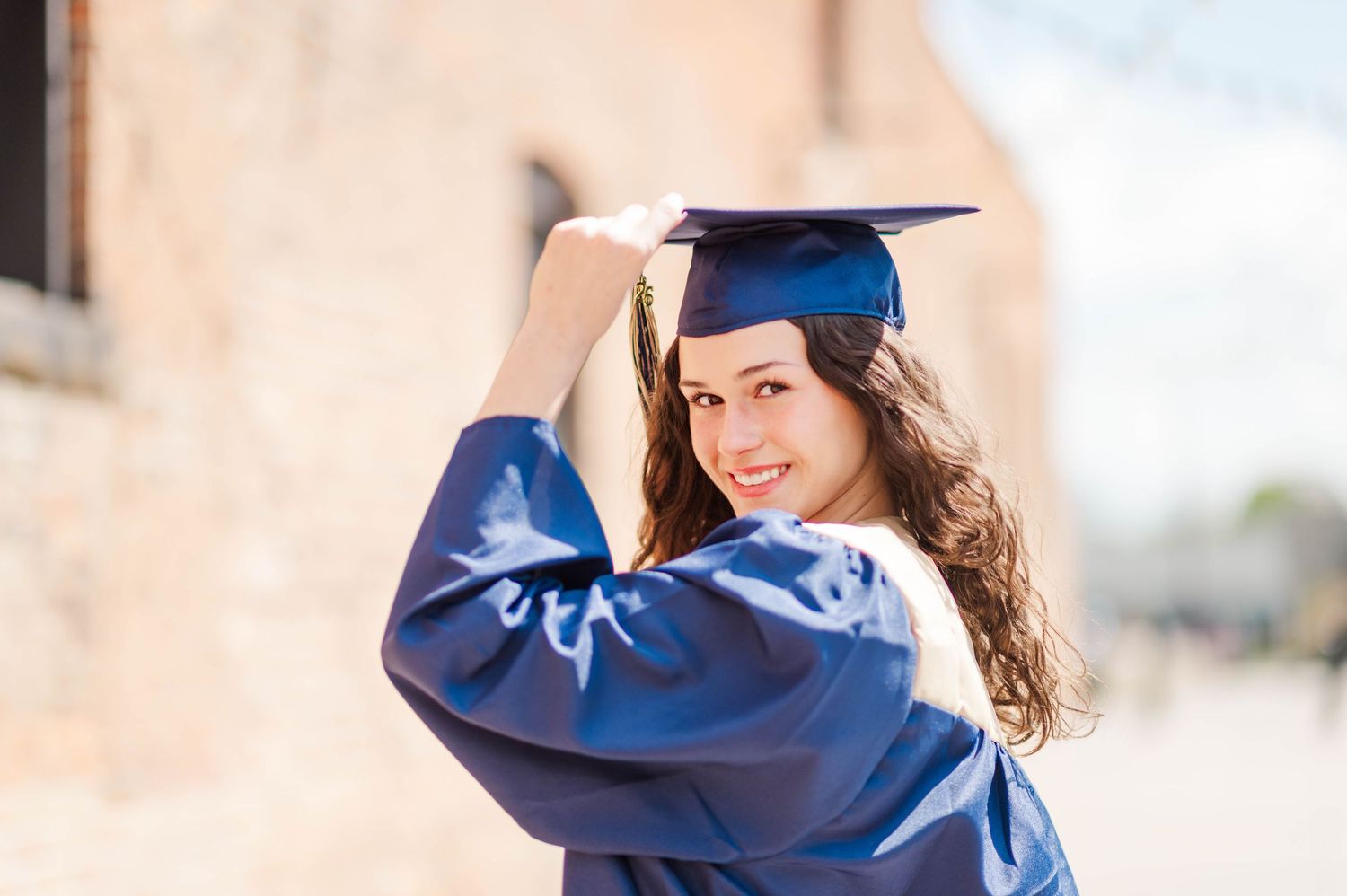 Smiling graduate in navy cap and gown poses outdoors adjusting mortarboard.