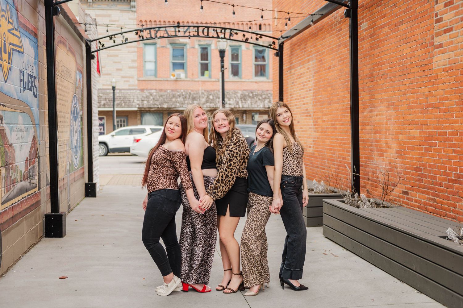 Group of five young women posing together under a decorative arch in a charming small town downtown area.