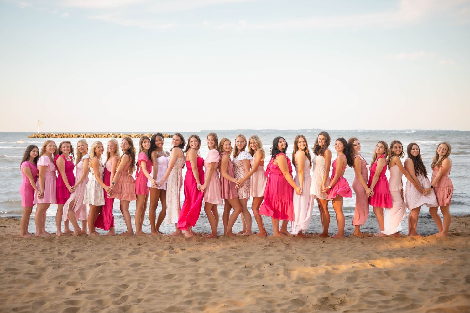 A group in pink and white dresses poses together on a beach at sunset.