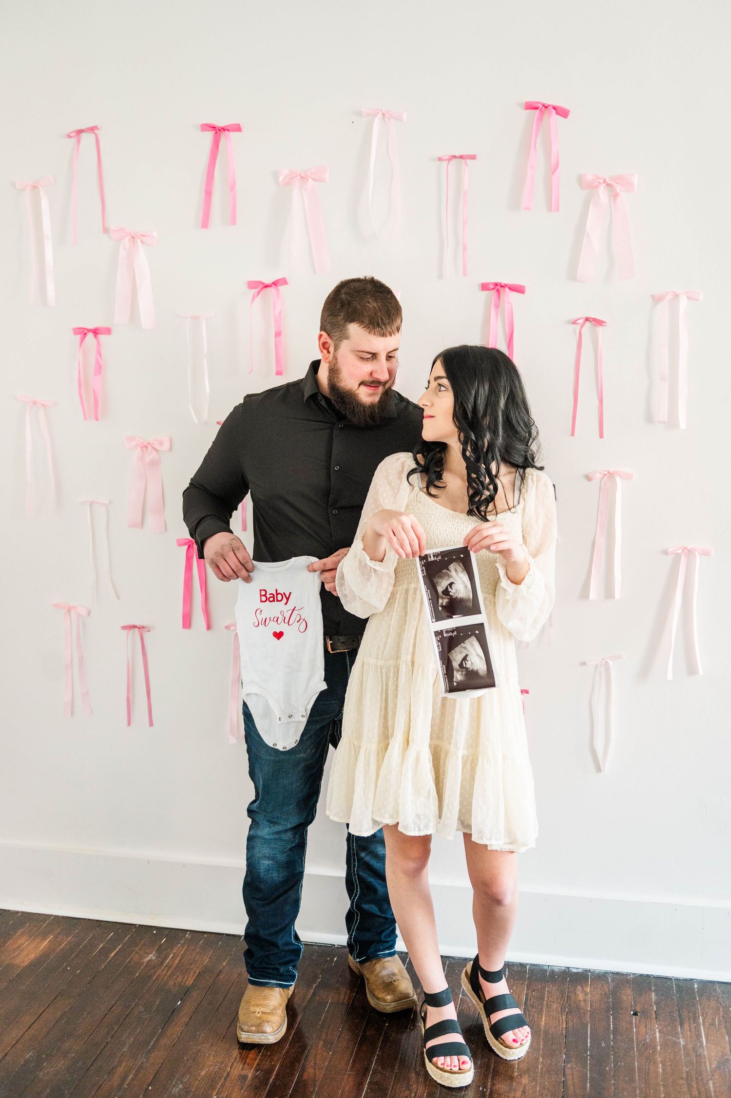 A couple shows excitement while displaying ultrasound photos against a wall with pink ribbons.