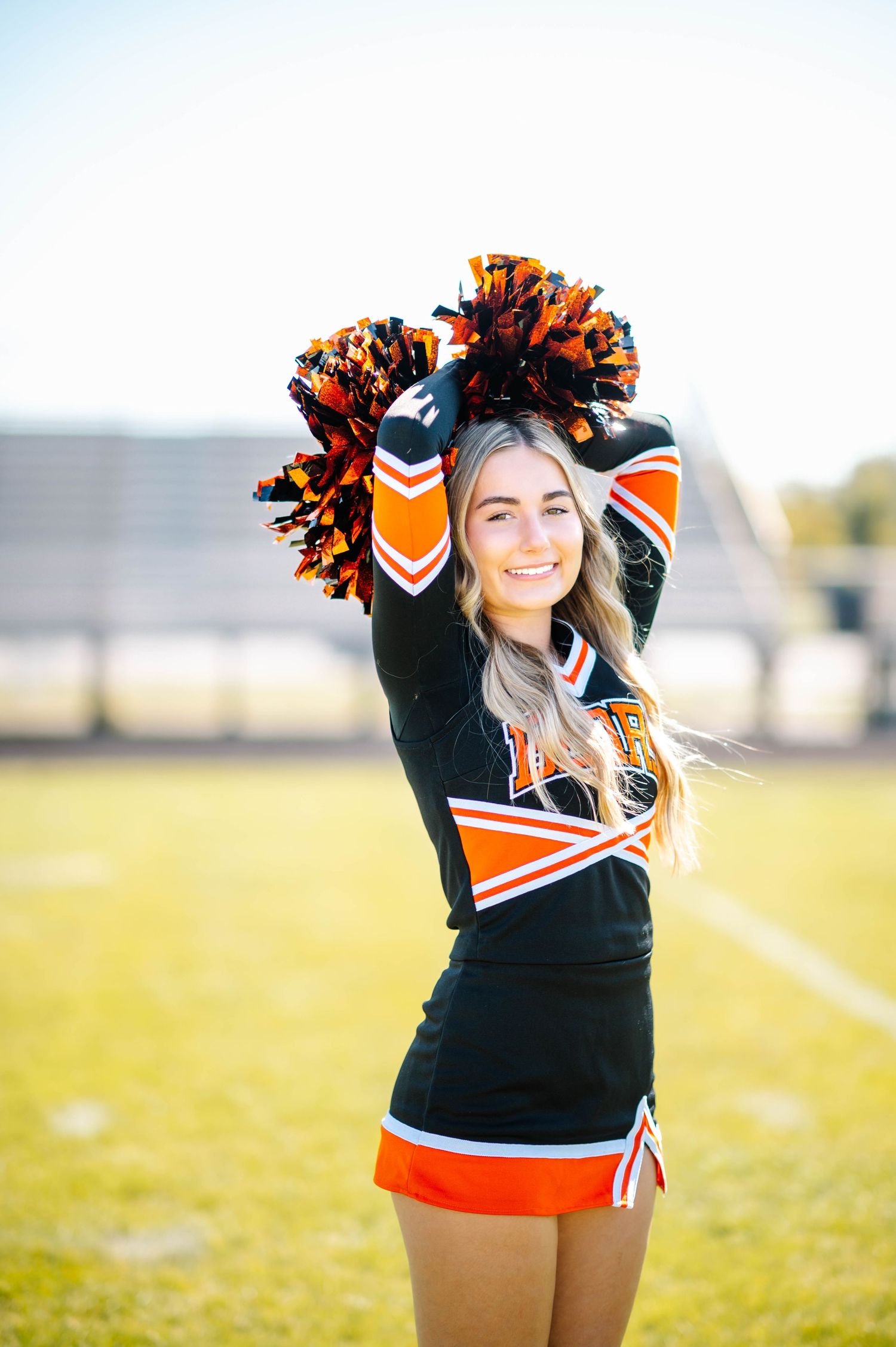 Cheerleader in black and orange uniform poses with pom poms on a sunny field.